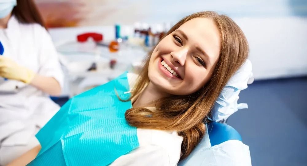 Woman Smiling in a Dental Chair; Dentist in Background, Blue Bib, Interior Setting — Tamworth Orthocare In Tamworth, NSW