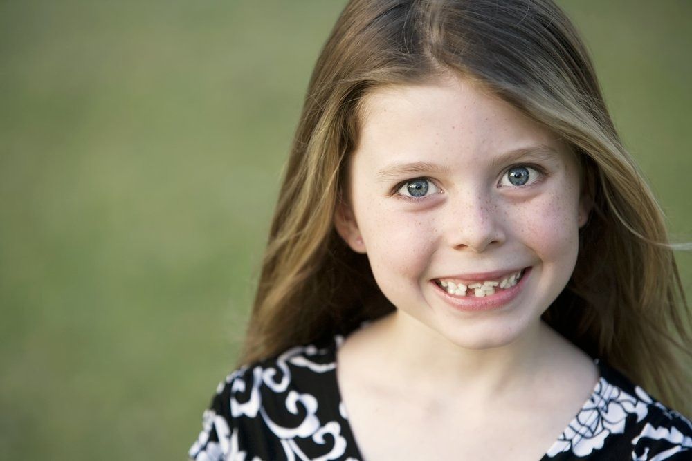 Girl smiling, missing front teeth, long blonde hair, black and white patterned top, green background.