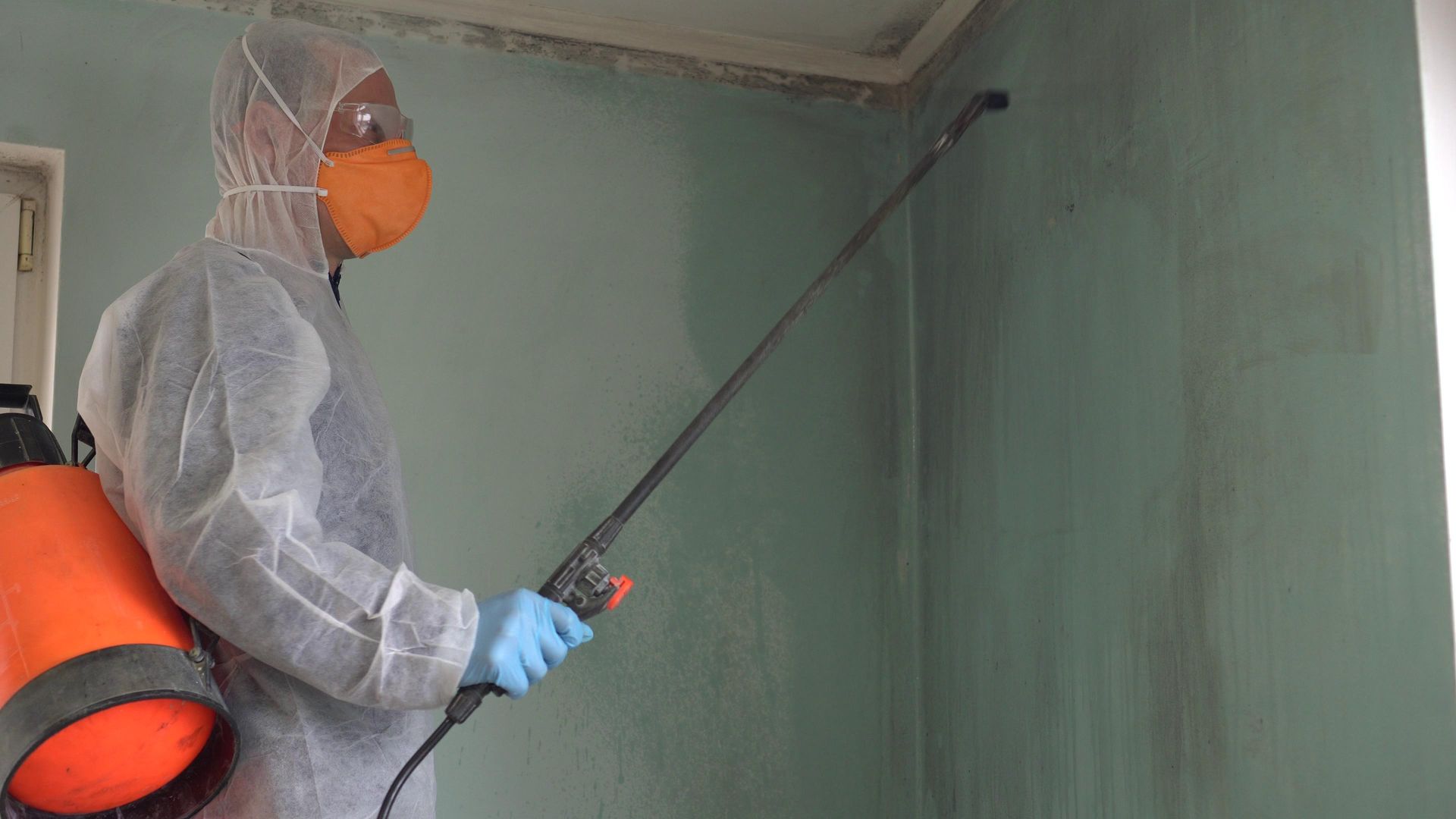 Person in protective suit spraying a wall, possibly for mold removal.