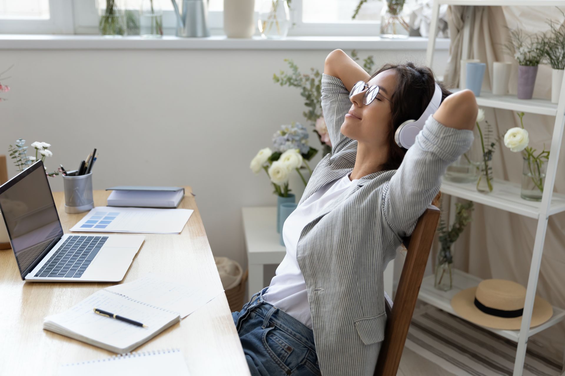 Woman in glasses with headphones, relaxing in chair at a desk with laptop, flowers, and notebook.