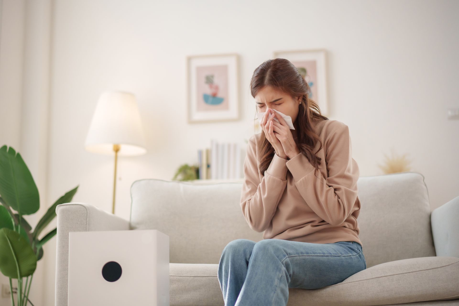 Woman in a beige sweater and jeans, sneezing into a tissue on a sofa, near an air purifier.