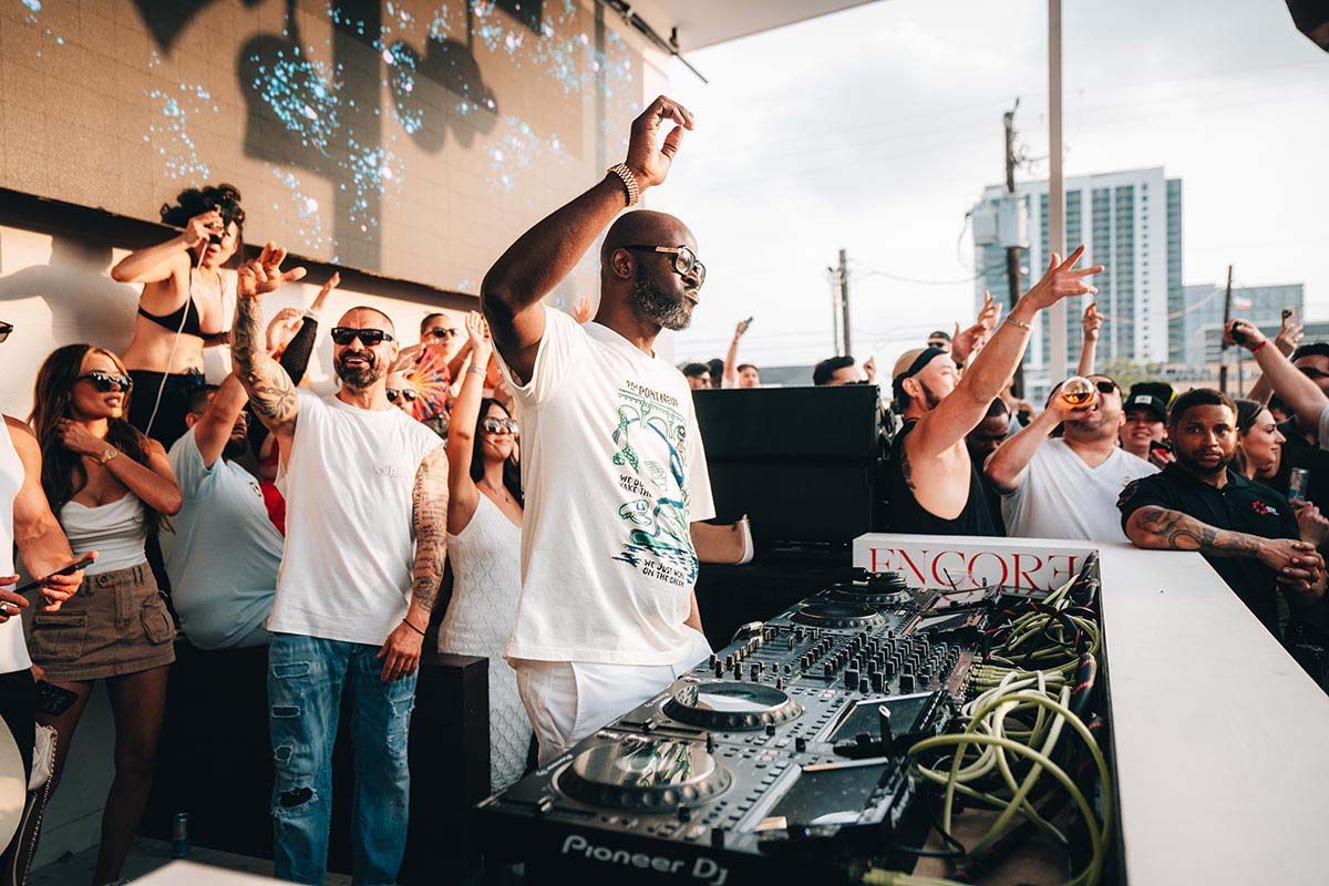 DJ at an outdoor party, raising arms. People in the crowd are cheering, daytime.