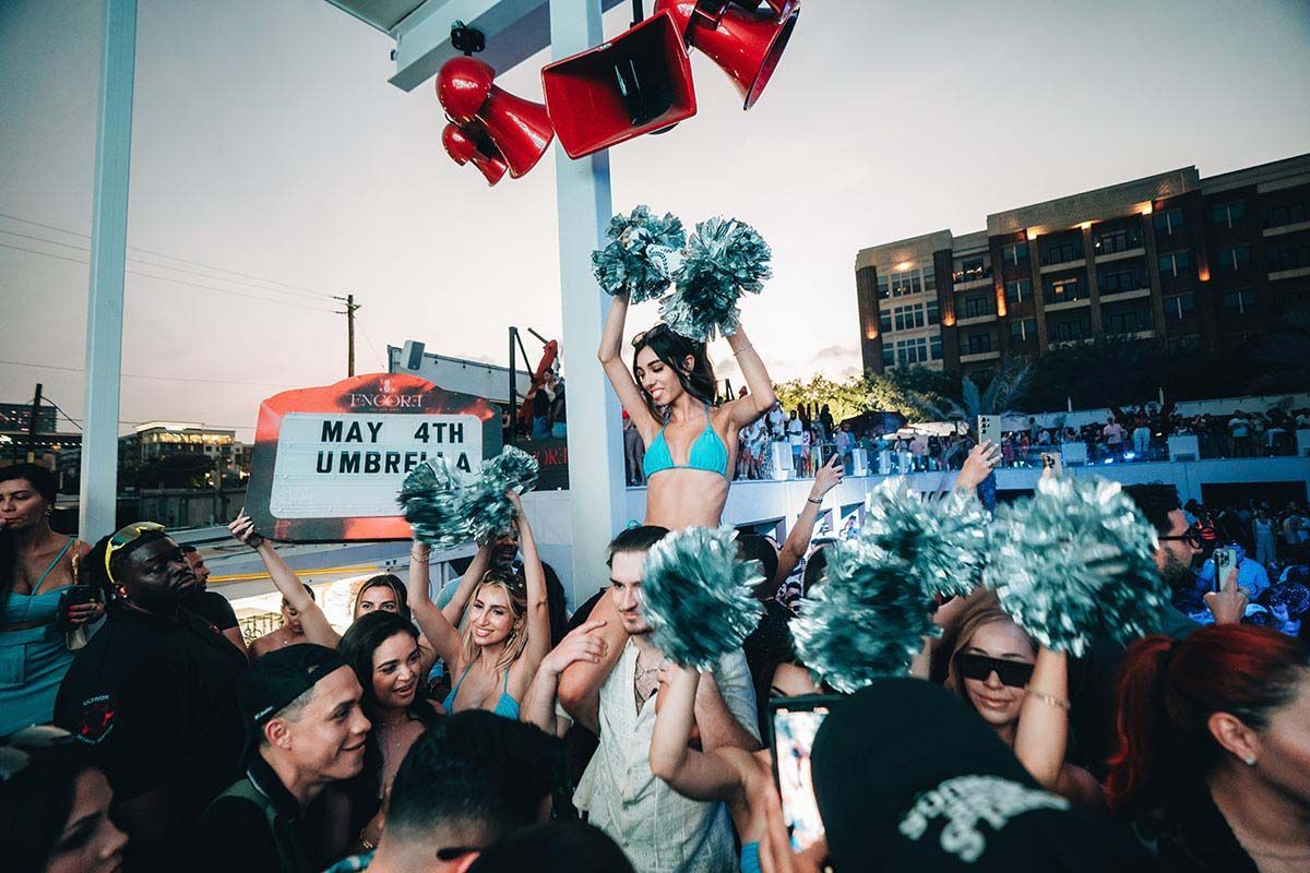 Party at a beach club with a woman on someone's shoulders holding pom-poms, and crowd.