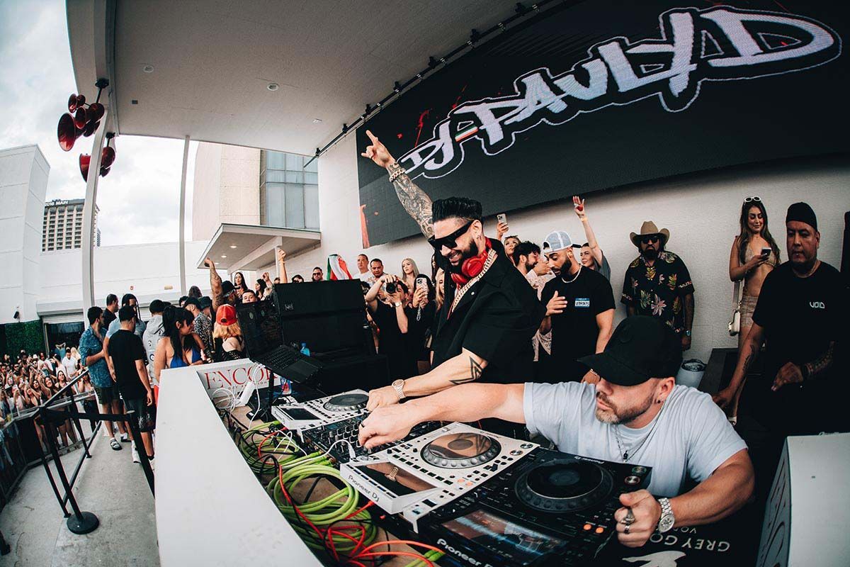 DJ Pauly D performing at an outdoor event. Crowd dances, DJ on turntables, logo banner overhead.