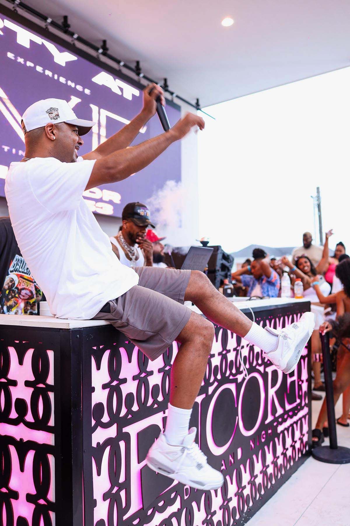 A performer in a white t-shirt and grey shorts sits on a stage edge, gesturing with a microphone during an event.