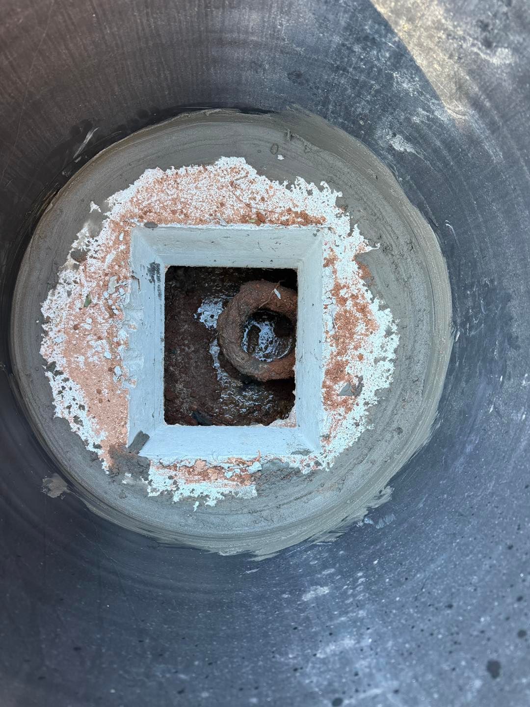 A square white drain set in circular gray concrete, with a dark, rusted metal pipe visible inside the opening.