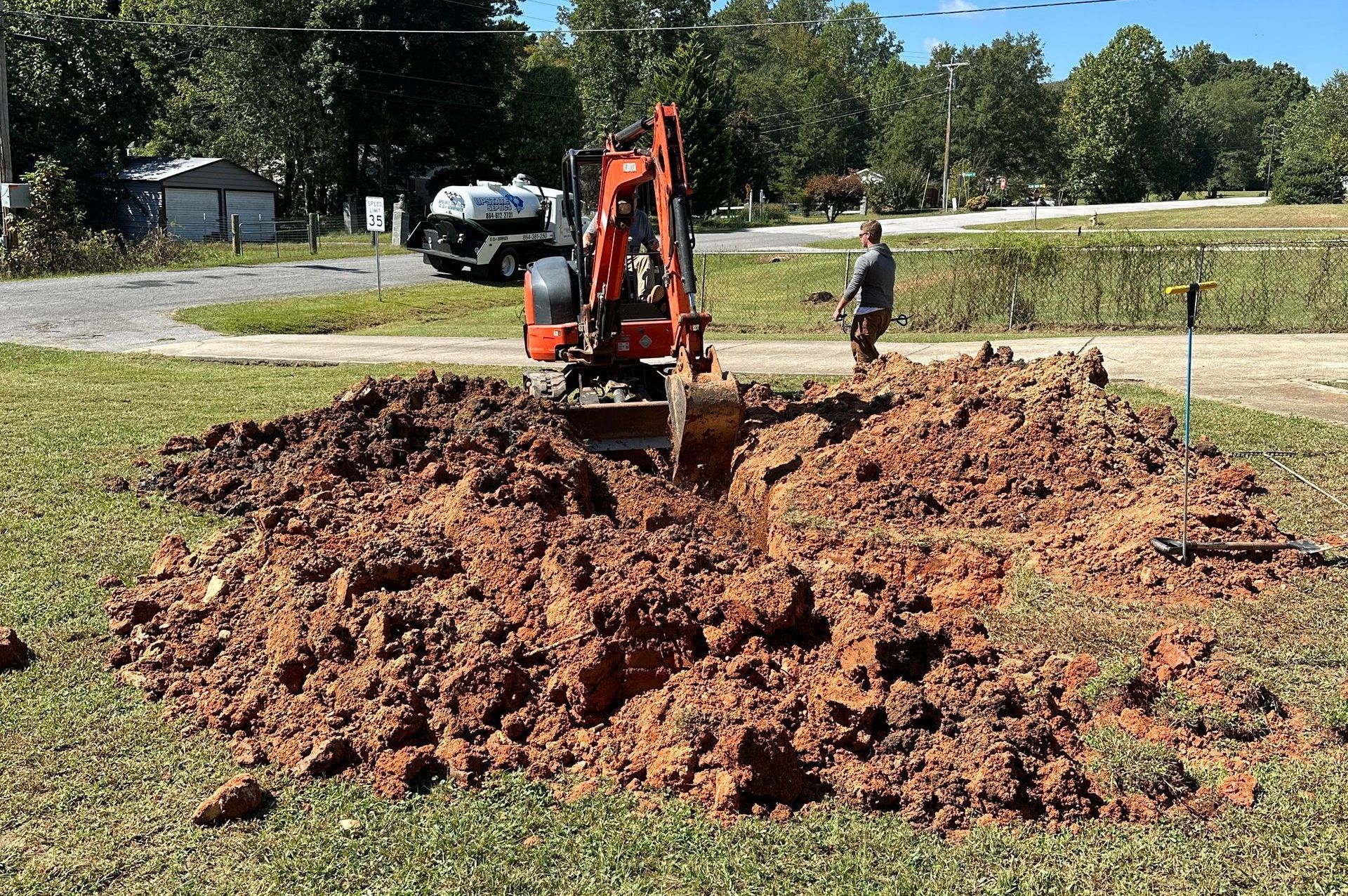 An orange excavator digs in a yard, pushing dirt into a large pile. A person stands nearby.