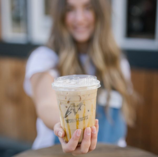 Woman holding out an iced coffee in a clear cup with caramel drizzle. Smiling outdoors.