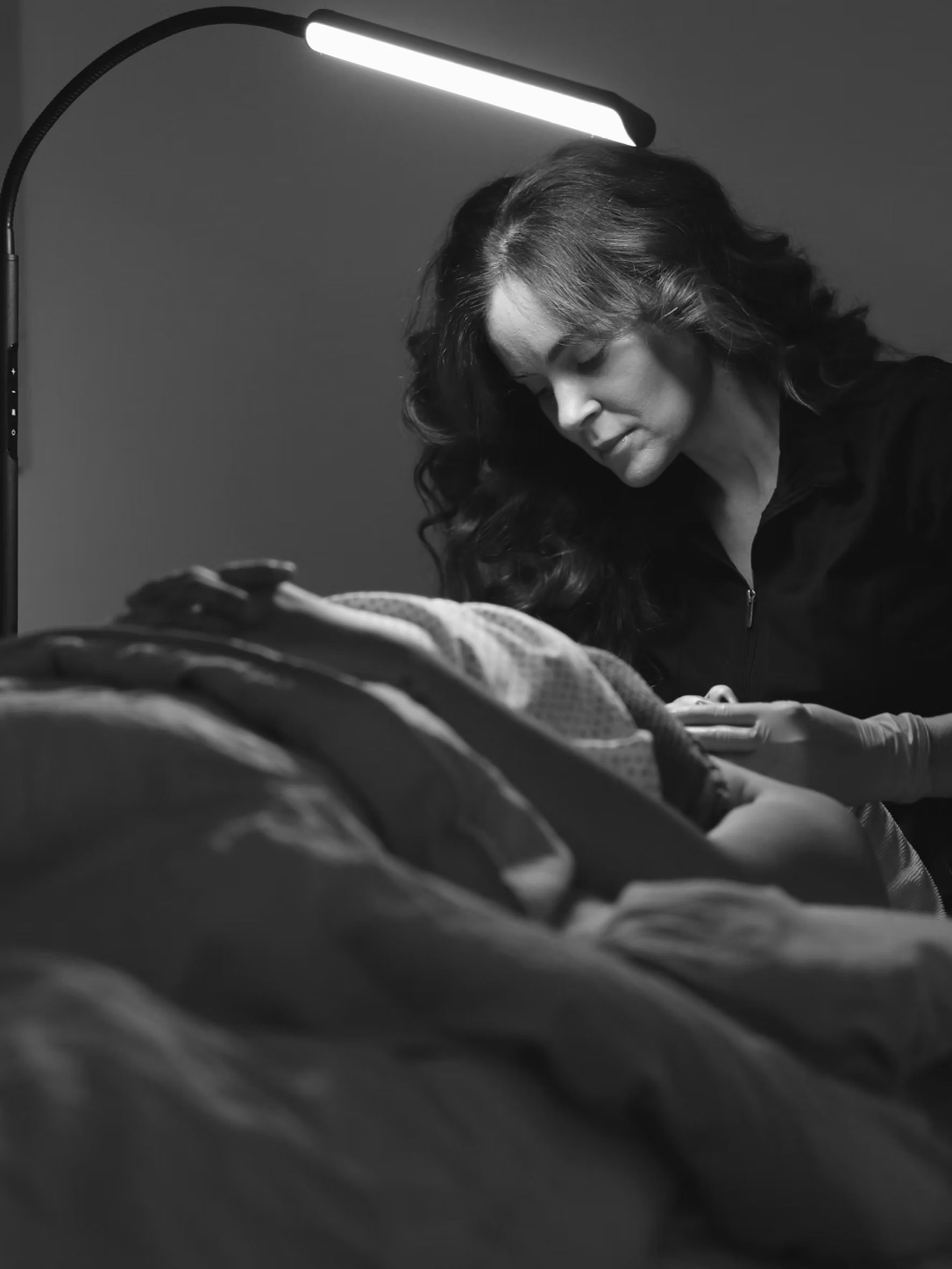 Person smiling beside a bed, reading under a bright lamp in a dim room