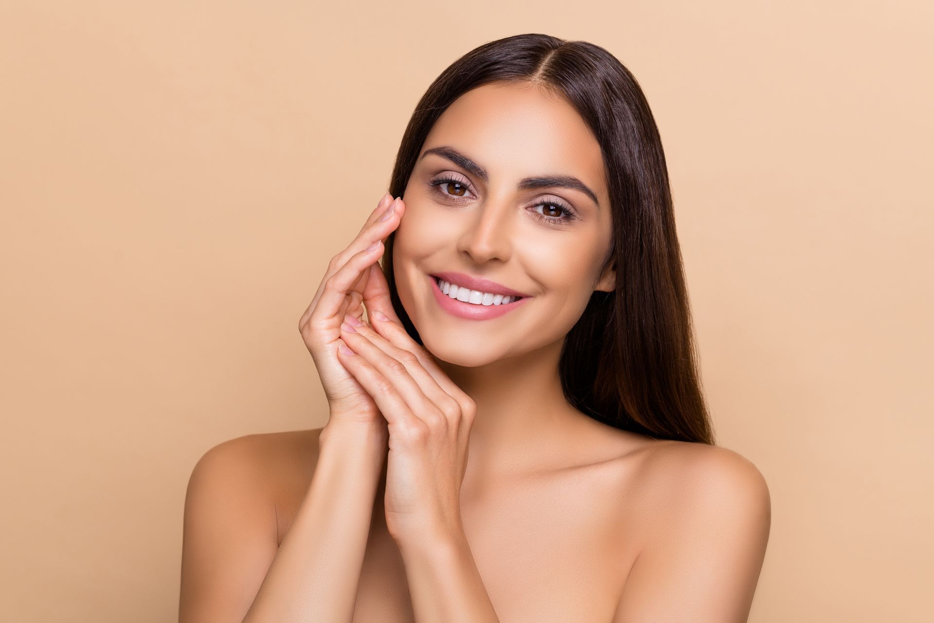 Smiling person with long dark hair posing against a beige background, hand resting on face