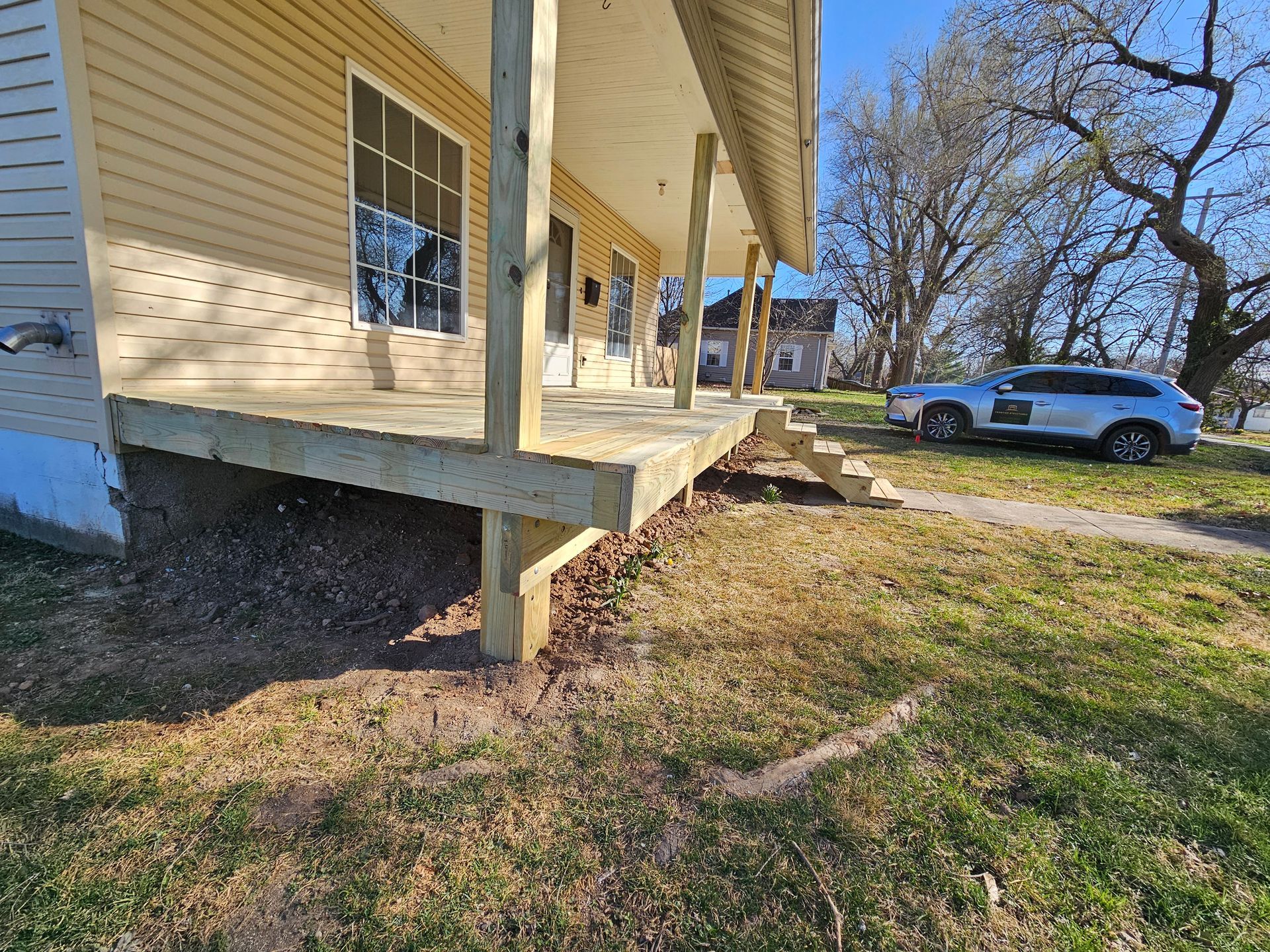 New wooden porch under construction on a house with beige siding. Brown grass and a car parked in the yard.