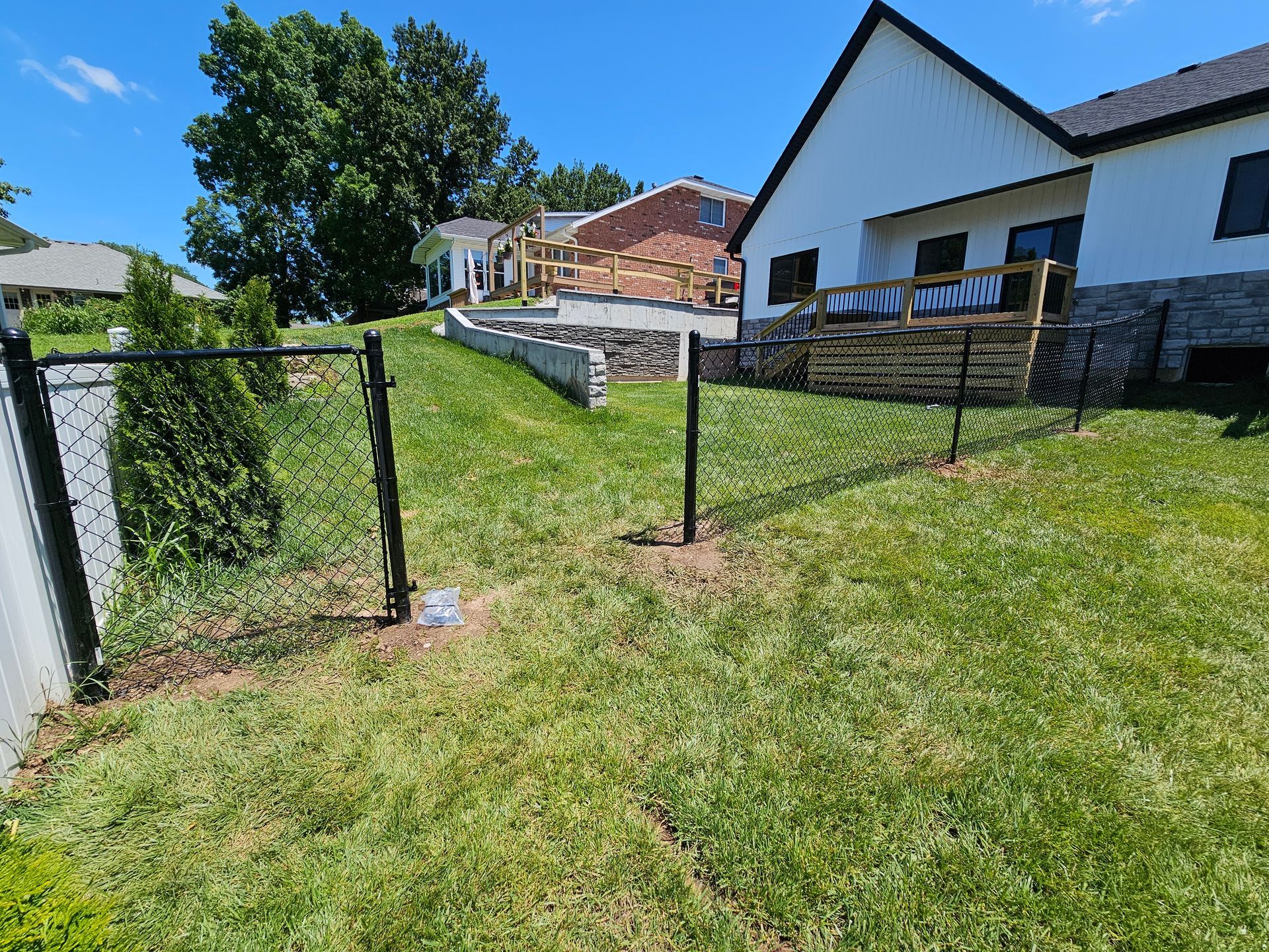 Black chain-link fence with an open gate in a grassy yard, near a white house and a raised garden bed.