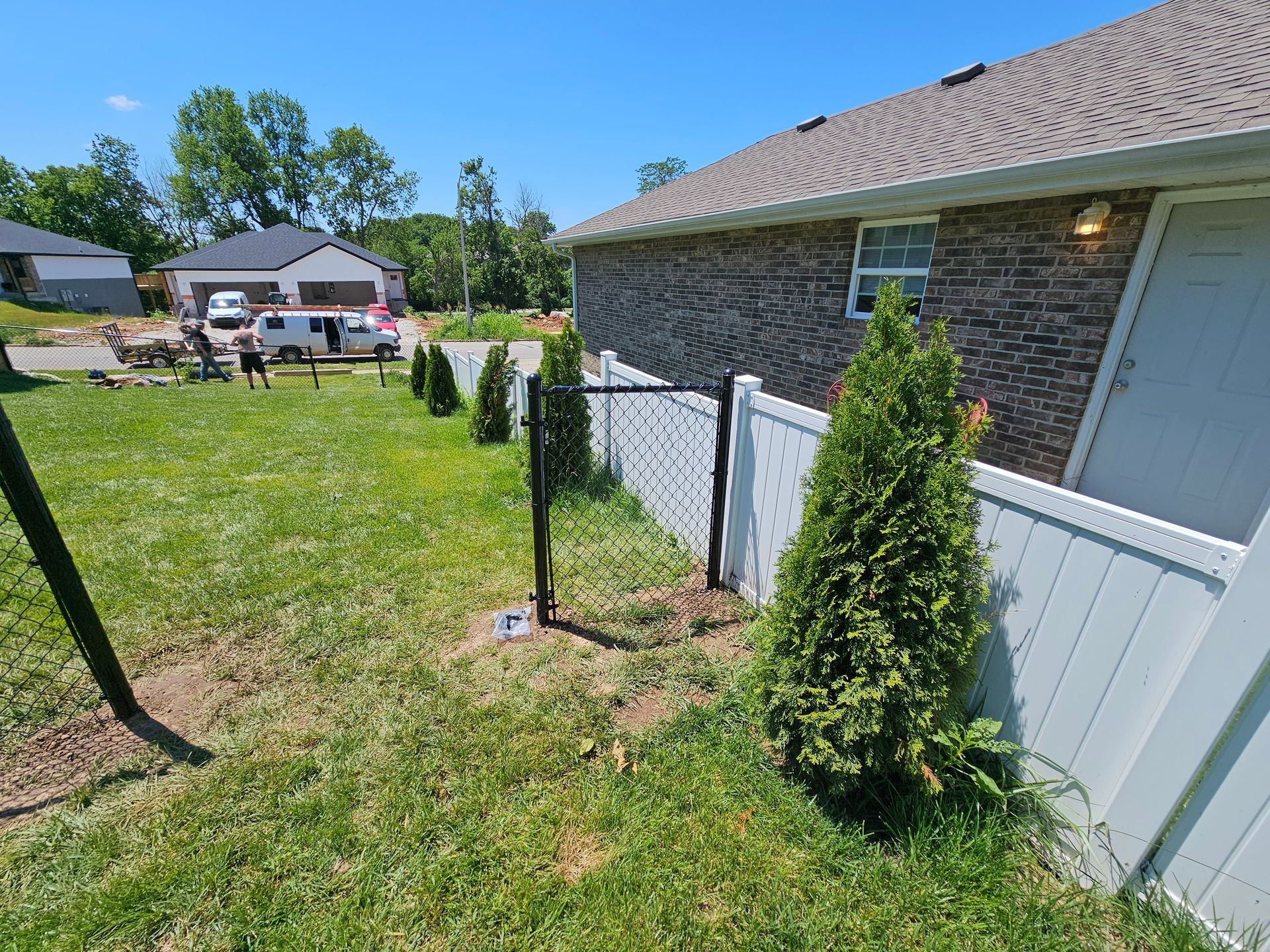 Green lawn with a white fence, black posts, and small evergreen trees alongside a brick house on a sunny day.