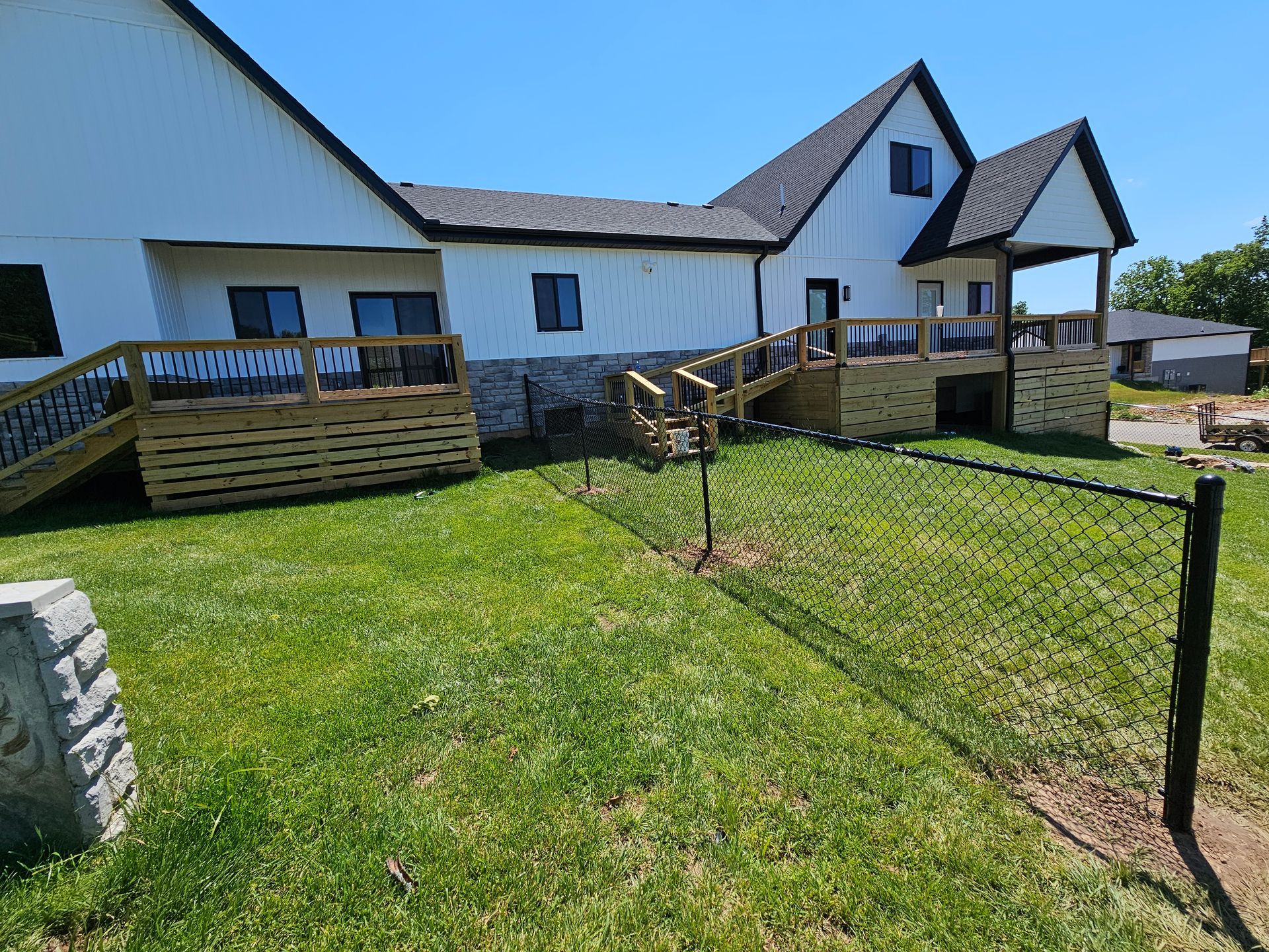 Exterior view of a white house with wooden decks and a chain-link fence on a green lawn.