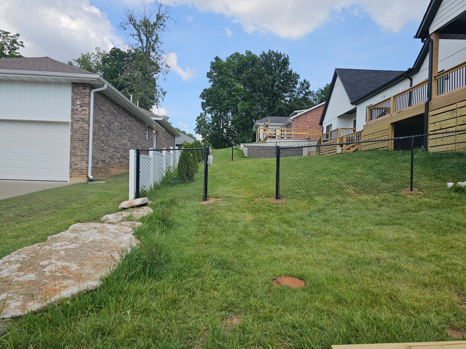 Chain-link fence on a grassy hill next to a house with a garage and a house with a deck under a cloudy sky.