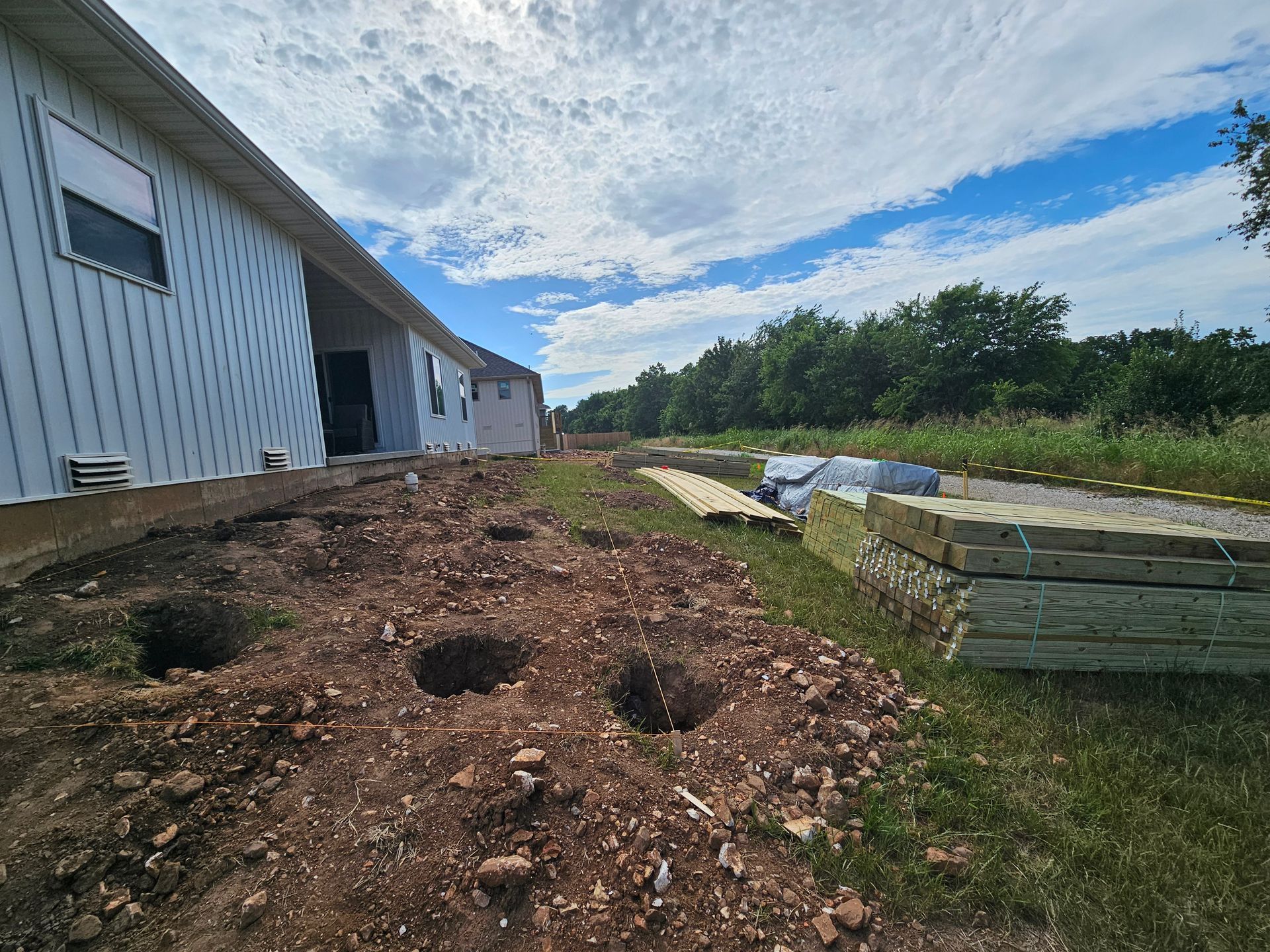 Construction site: building with white siding, holes in dirt, and stacked materials under blue sky.