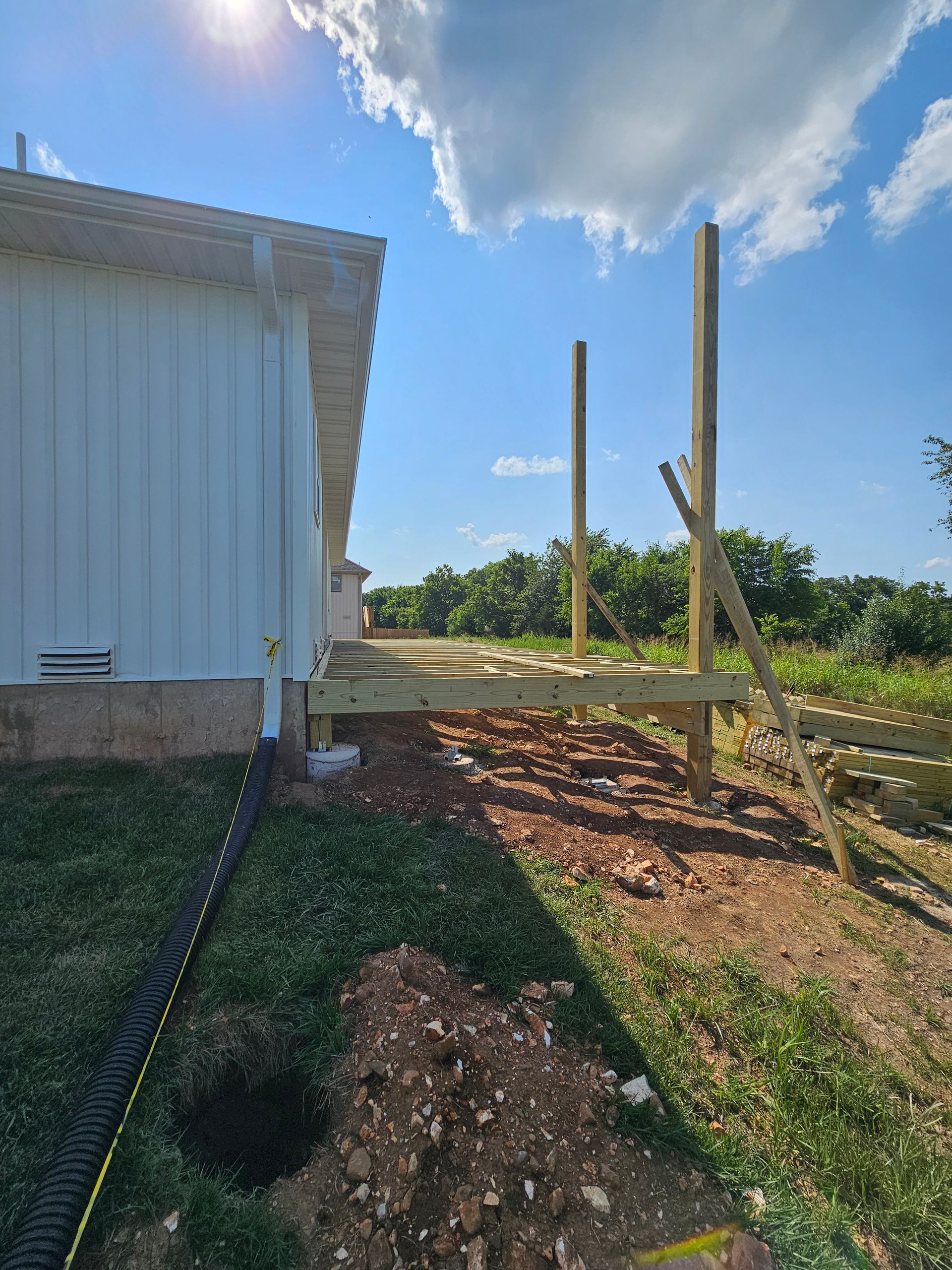 Construction of a wooden deck next to a white building; two support posts stand; blue sky and clouds.