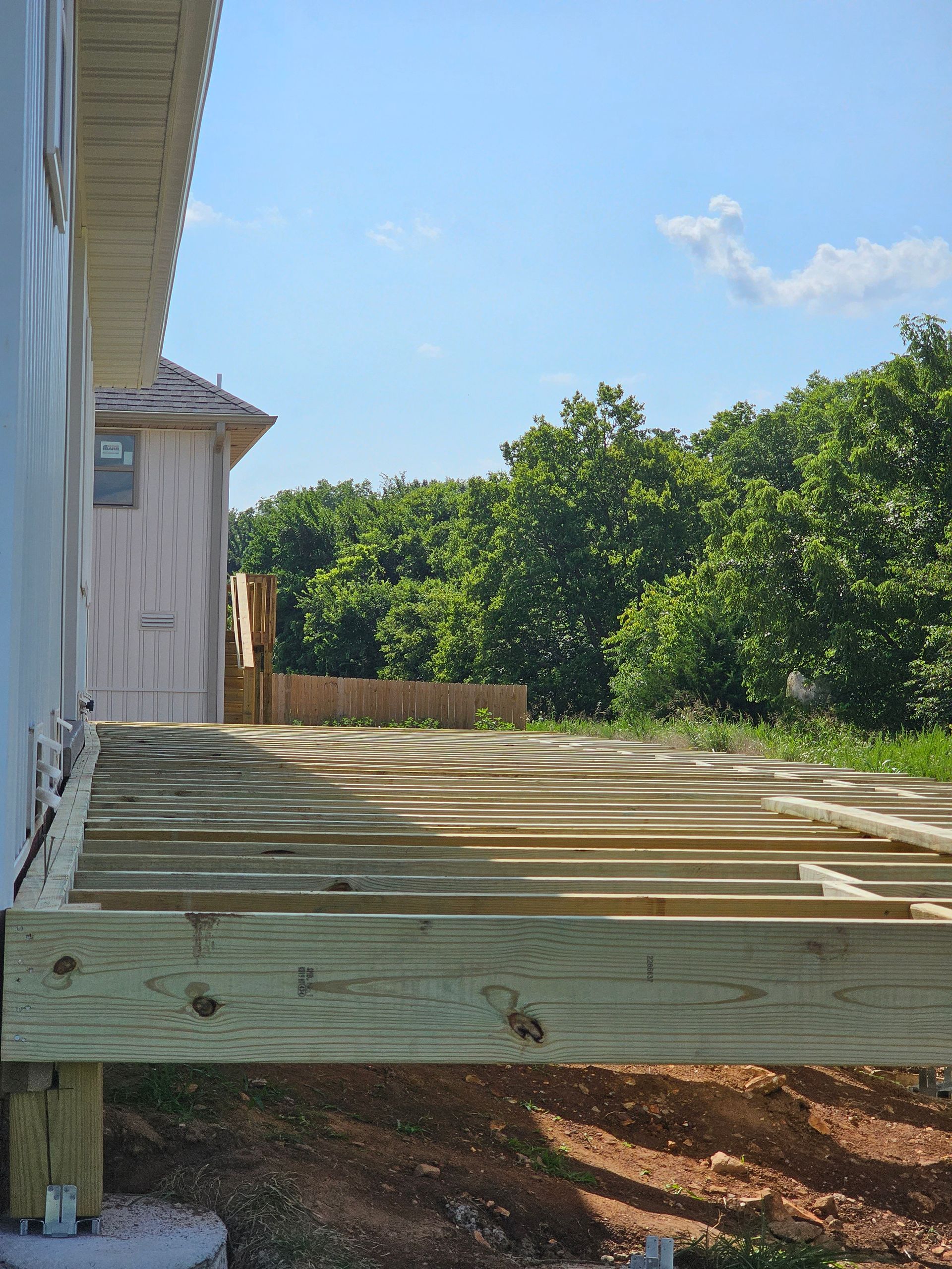 Wooden deck under construction next to a light-colored building, with trees and a blue sky in the background.