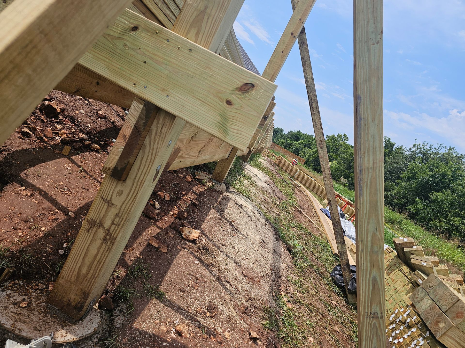 Wooden support structure being built on a hillside, with blue sky and trees in the background.