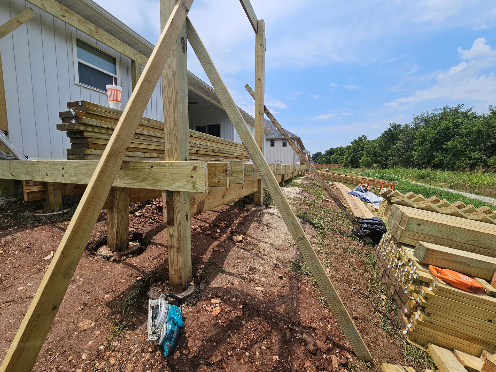 Deck construction: wood posts, framing, and stacked boards along a house and dirt path under a blue sky.