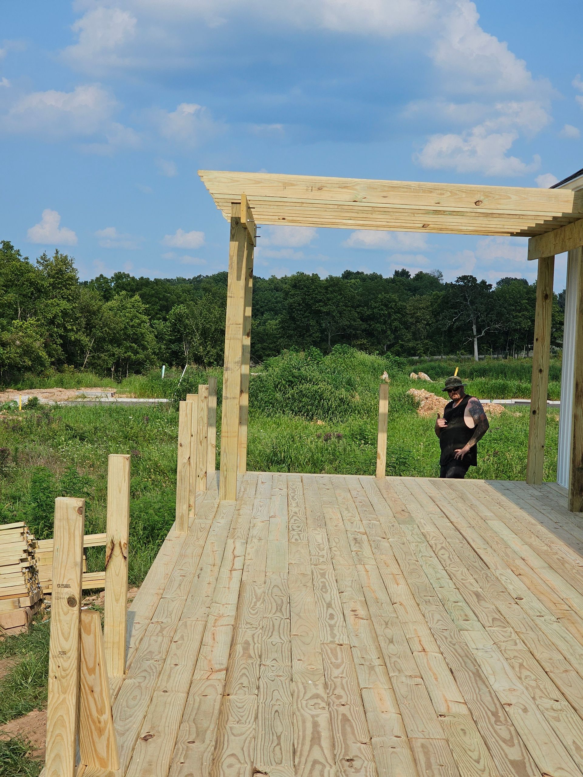 Wooden deck and pergola under construction with person standing in the background. Green trees and blue sky.