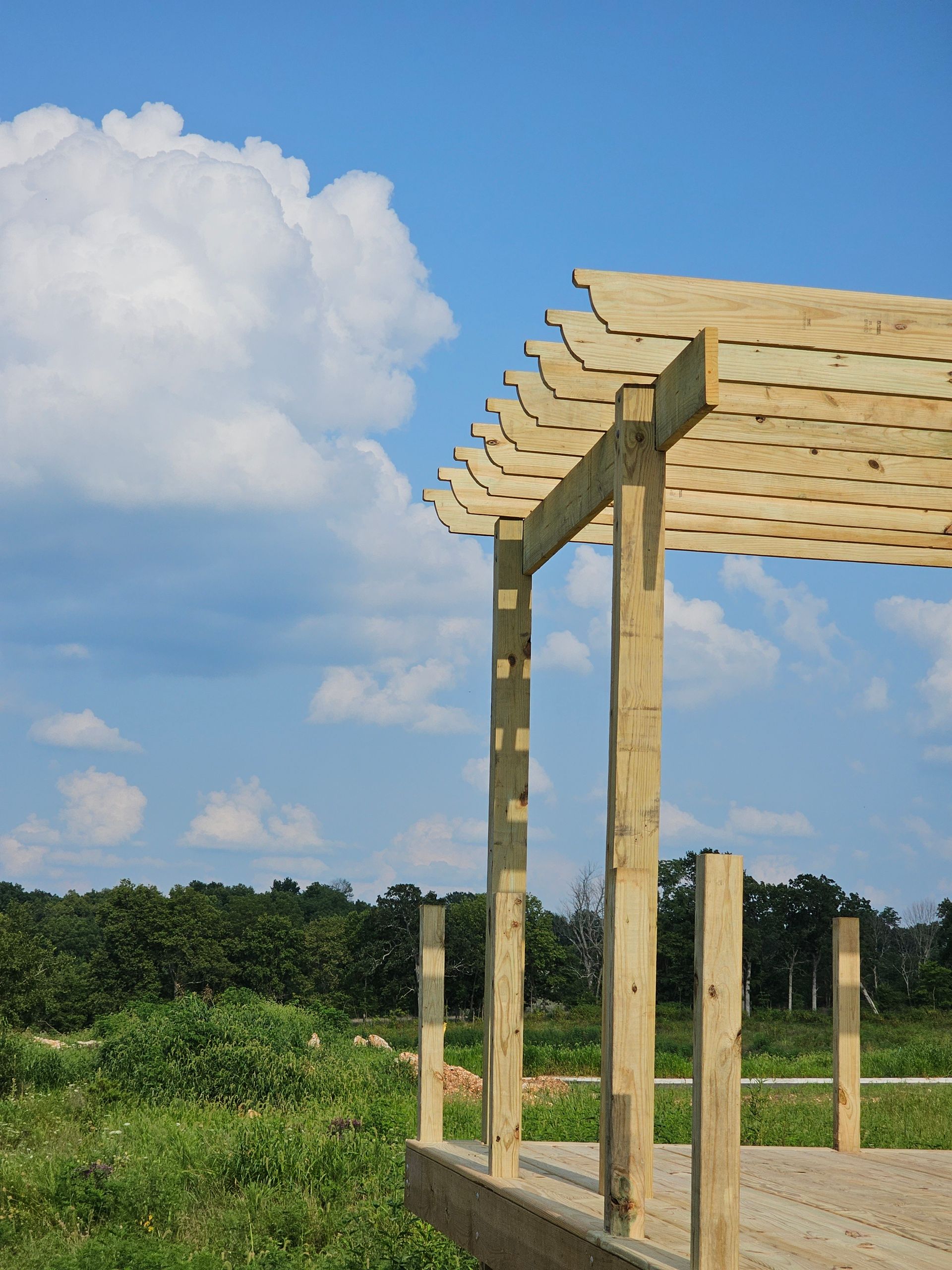 Wooden pergola under construction against a blue sky with puffy clouds; a green field in the foreground.