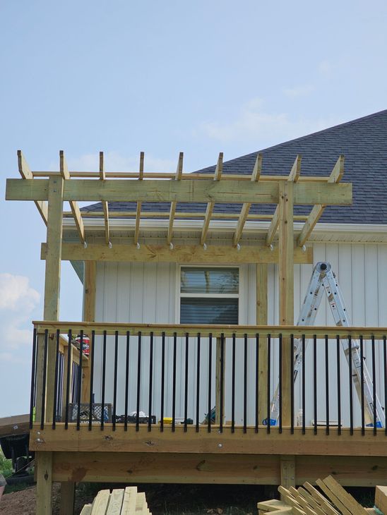 Wooden deck with railing and partially built pergola next to a white house with a dark roof.
