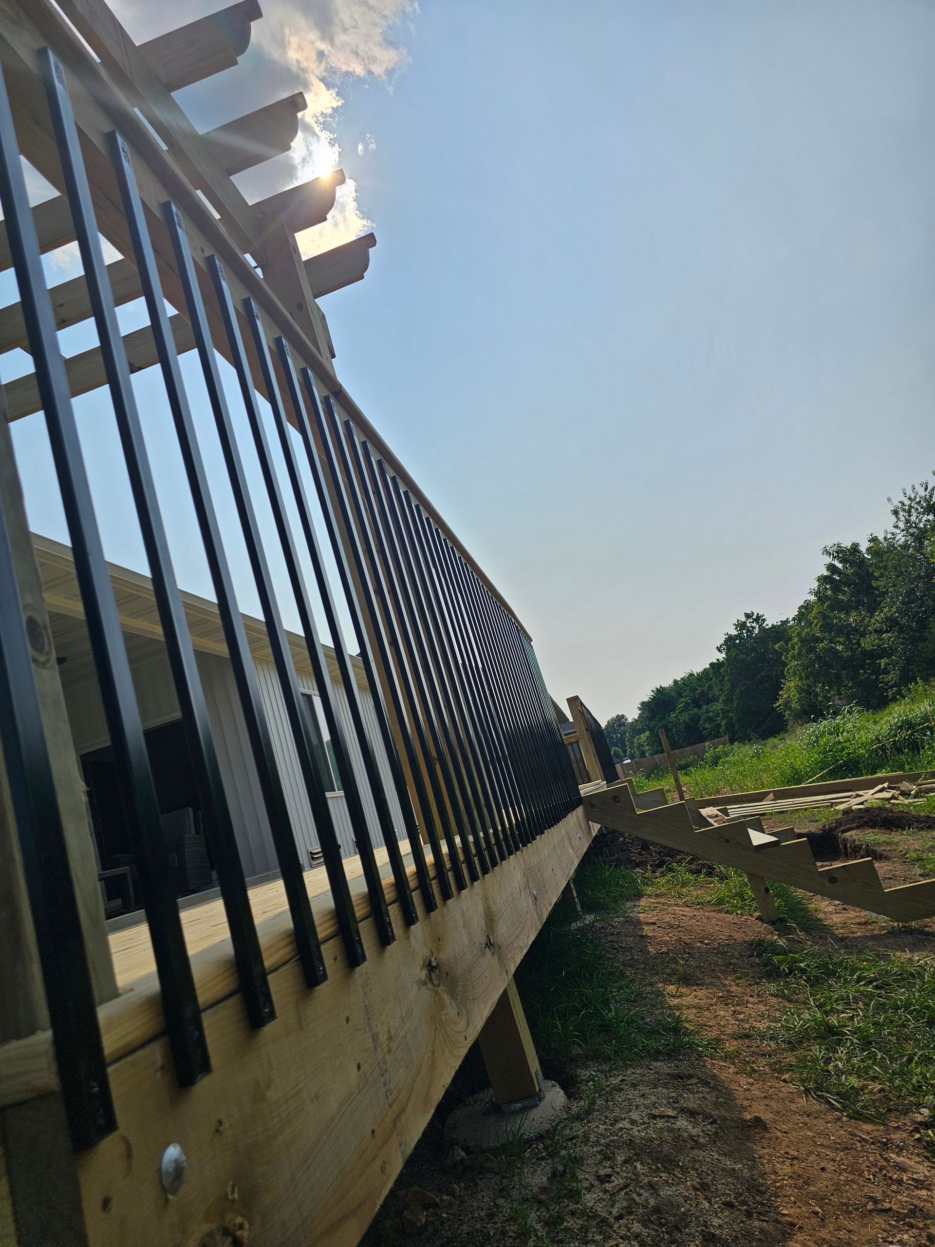 Wooden deck with black railing under a blue sky, sunlight peeking through pergola.