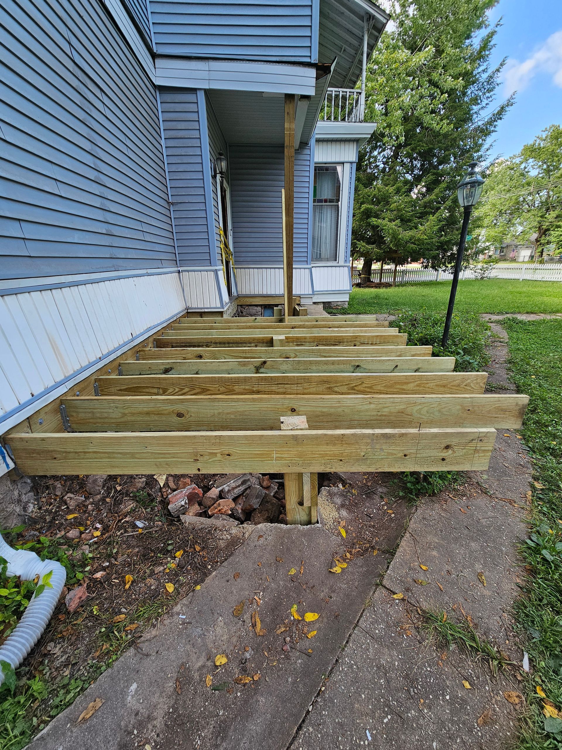 Wooden deck under construction with exposed beams. Steps lead to a blue house entrance.