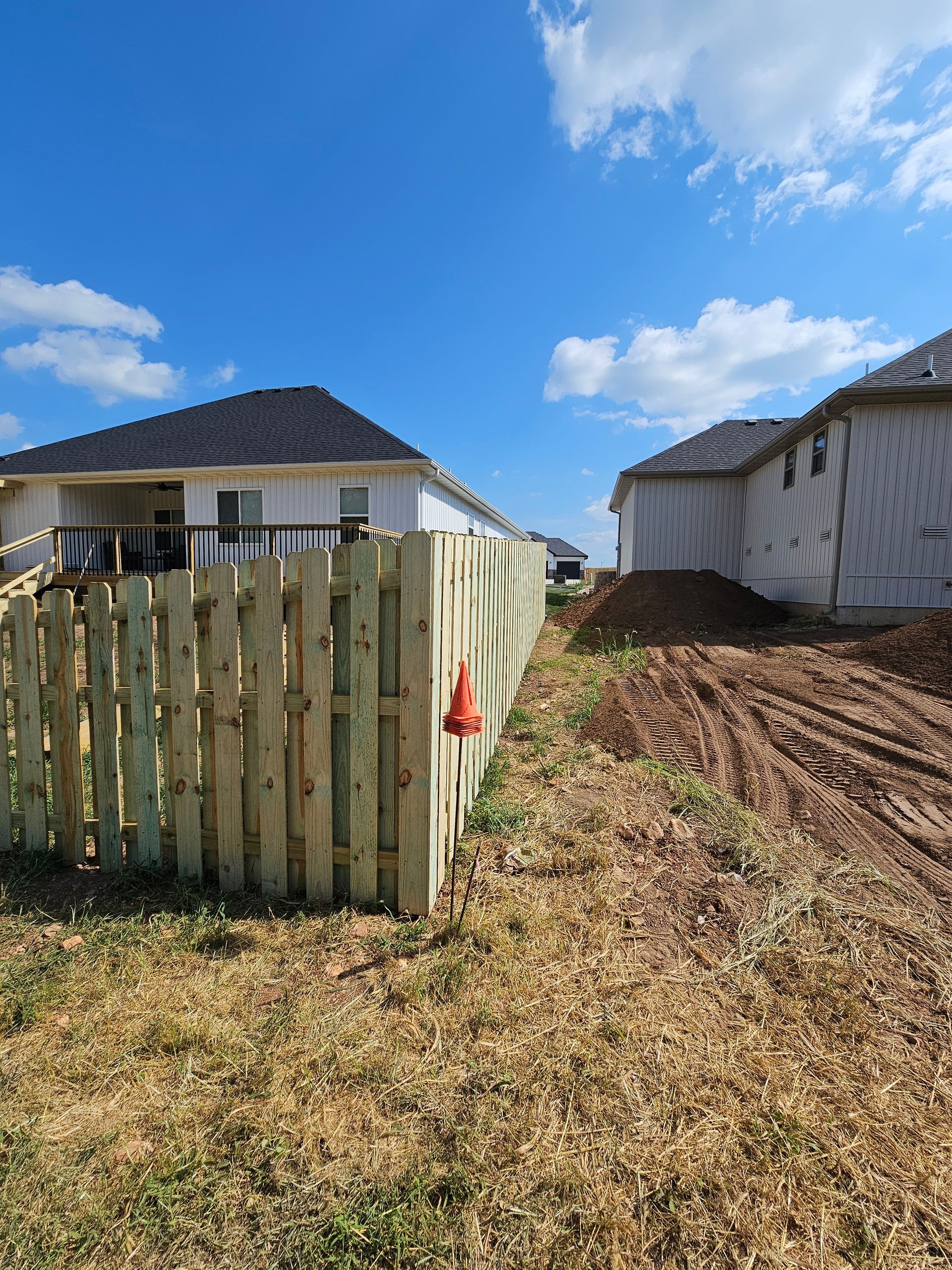 Wooden fence bordering a grassy yard with dirt pile next to a house under construction; blue sky.
