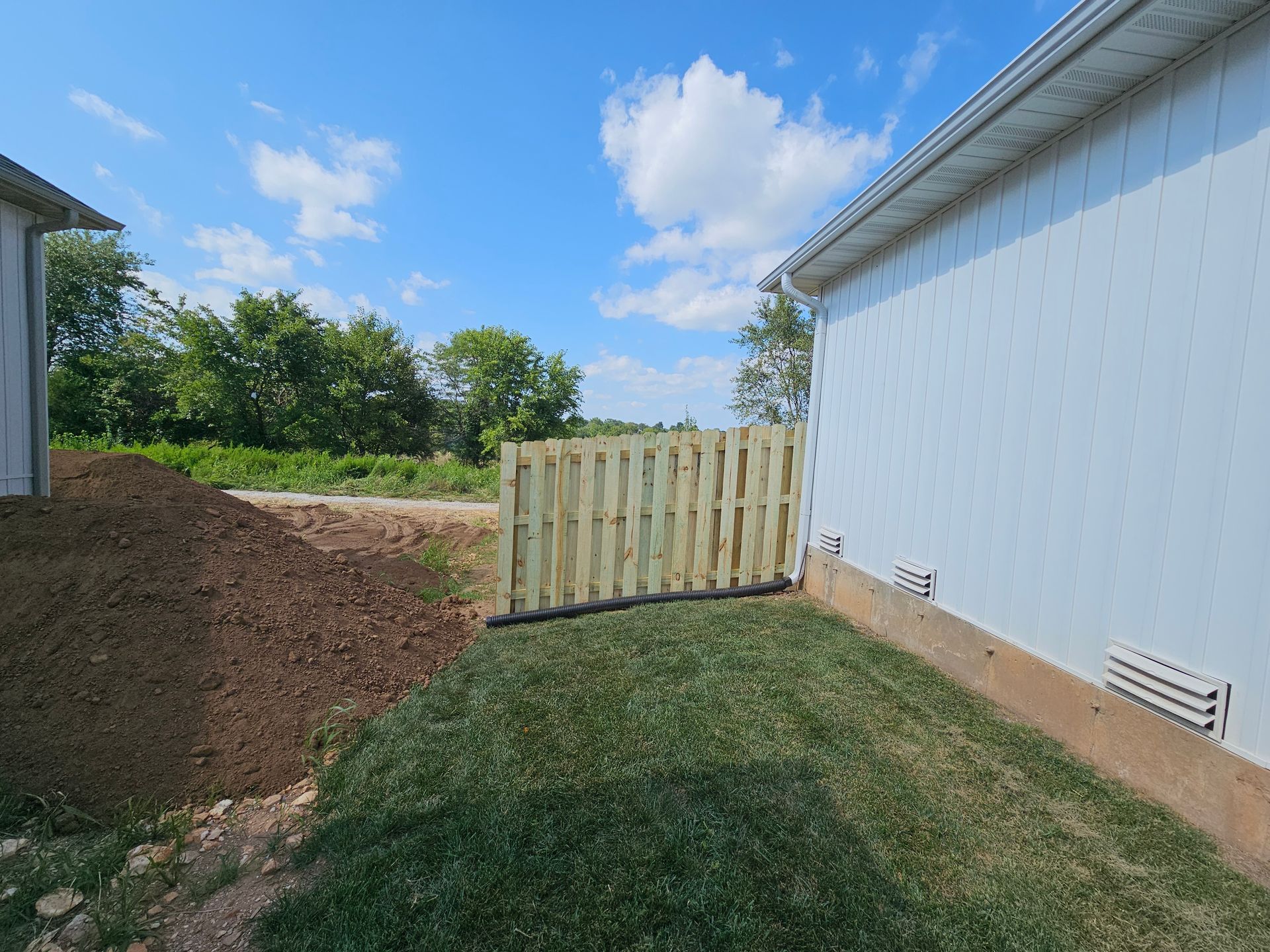 A wooden fence between a house and a dirt pile, with grass and blue sky.