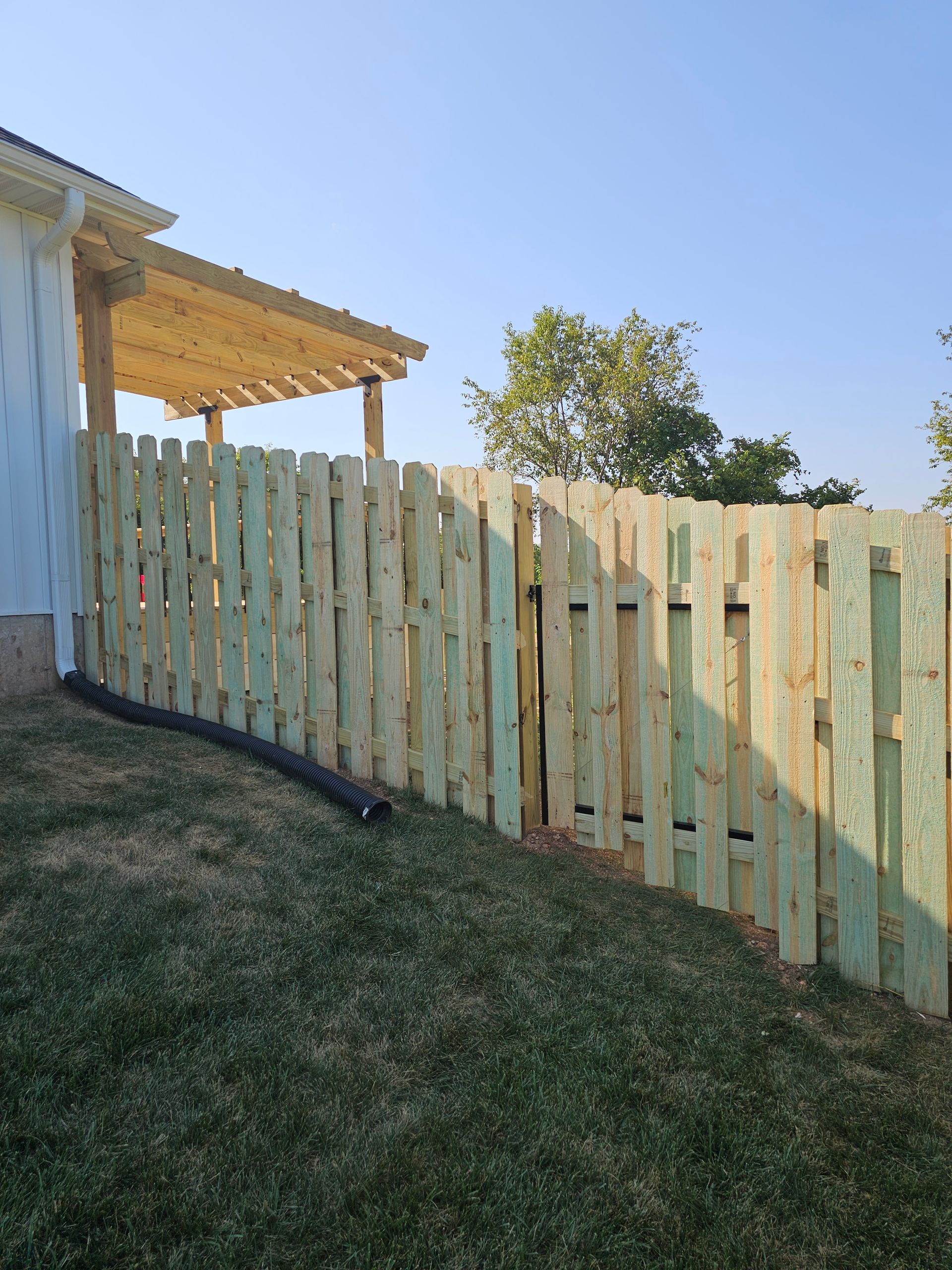 Wooden fence alongside a building with a pergola, on a grassy hillside under a blue sky.