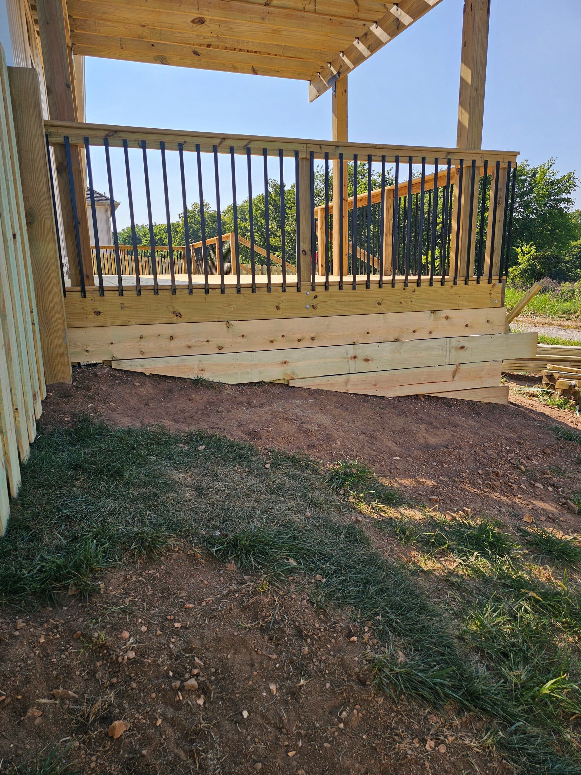 Wooden deck with railing, constructed over a sloped dirt and plant bed, with a pergola roof overhead.