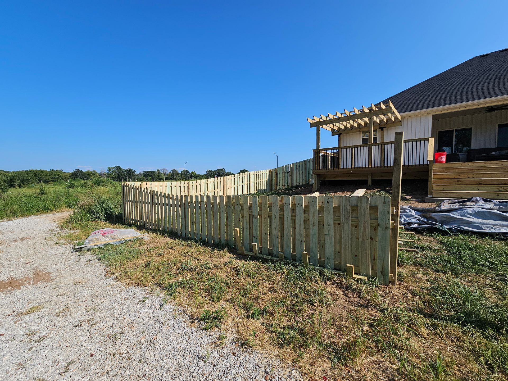 Wooden fence curves along a yard, next to gravel and a house with a deck. Blue sky overhead.