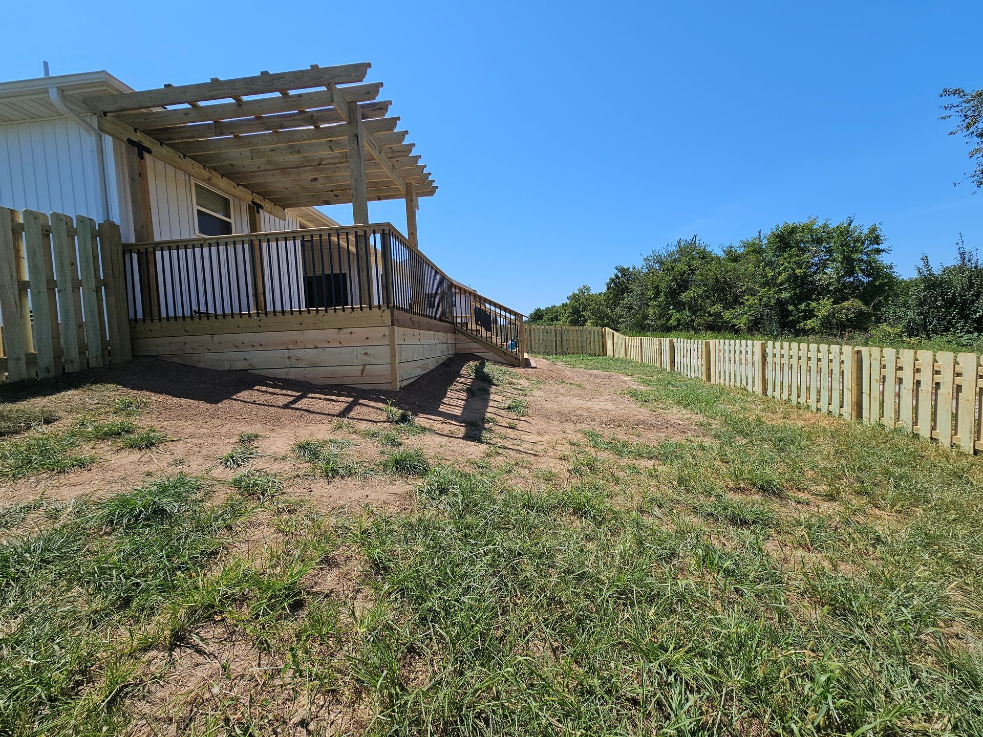 Backyard with a wooden deck, fence, and a pergola. Green grass and blue sky.