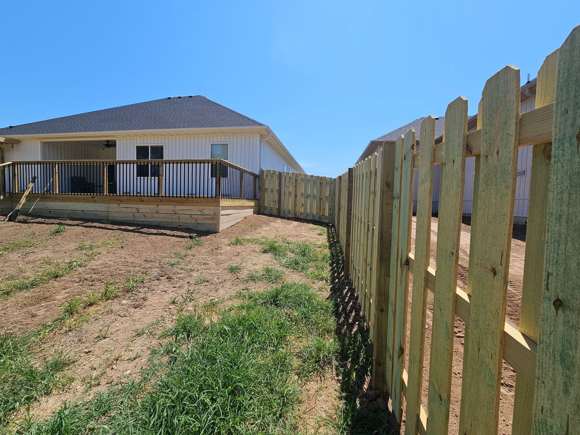Wooden picket fence bordering a yard with a house in the background on a sunny day.