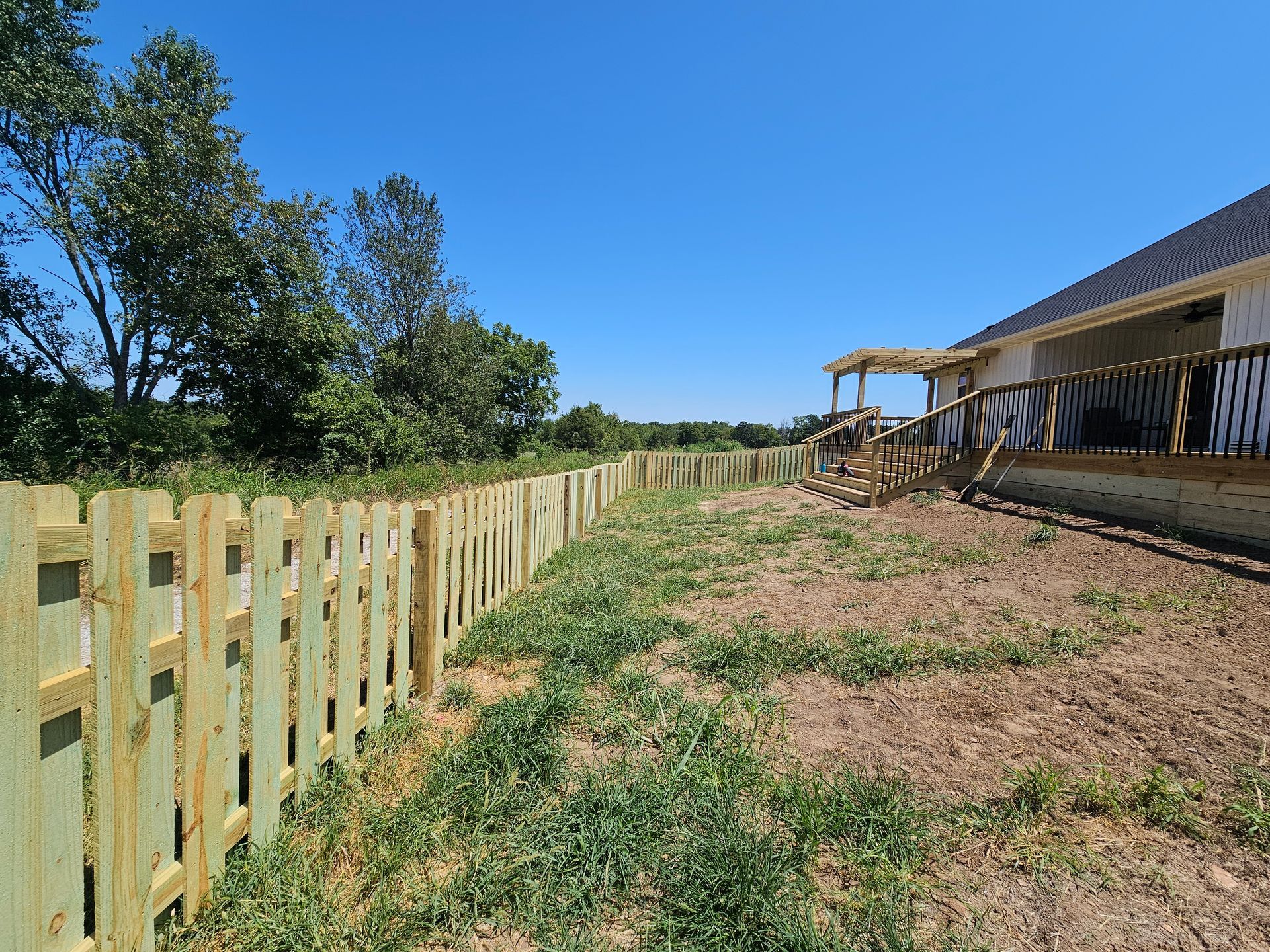 Wooden picket fence along grassy yard, next to a deck under blue sky.