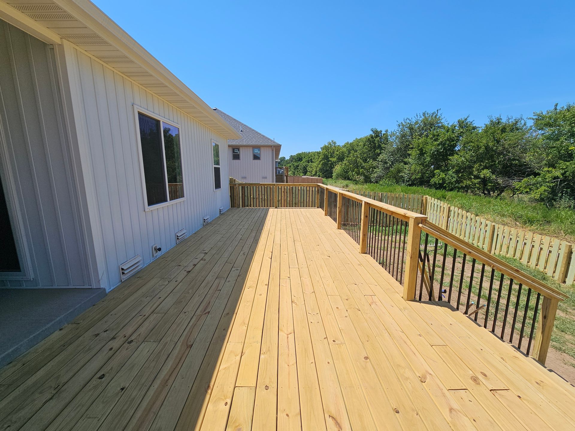 Wooden deck on a sunny day next to a white house with black window frames, overlooking a yard.