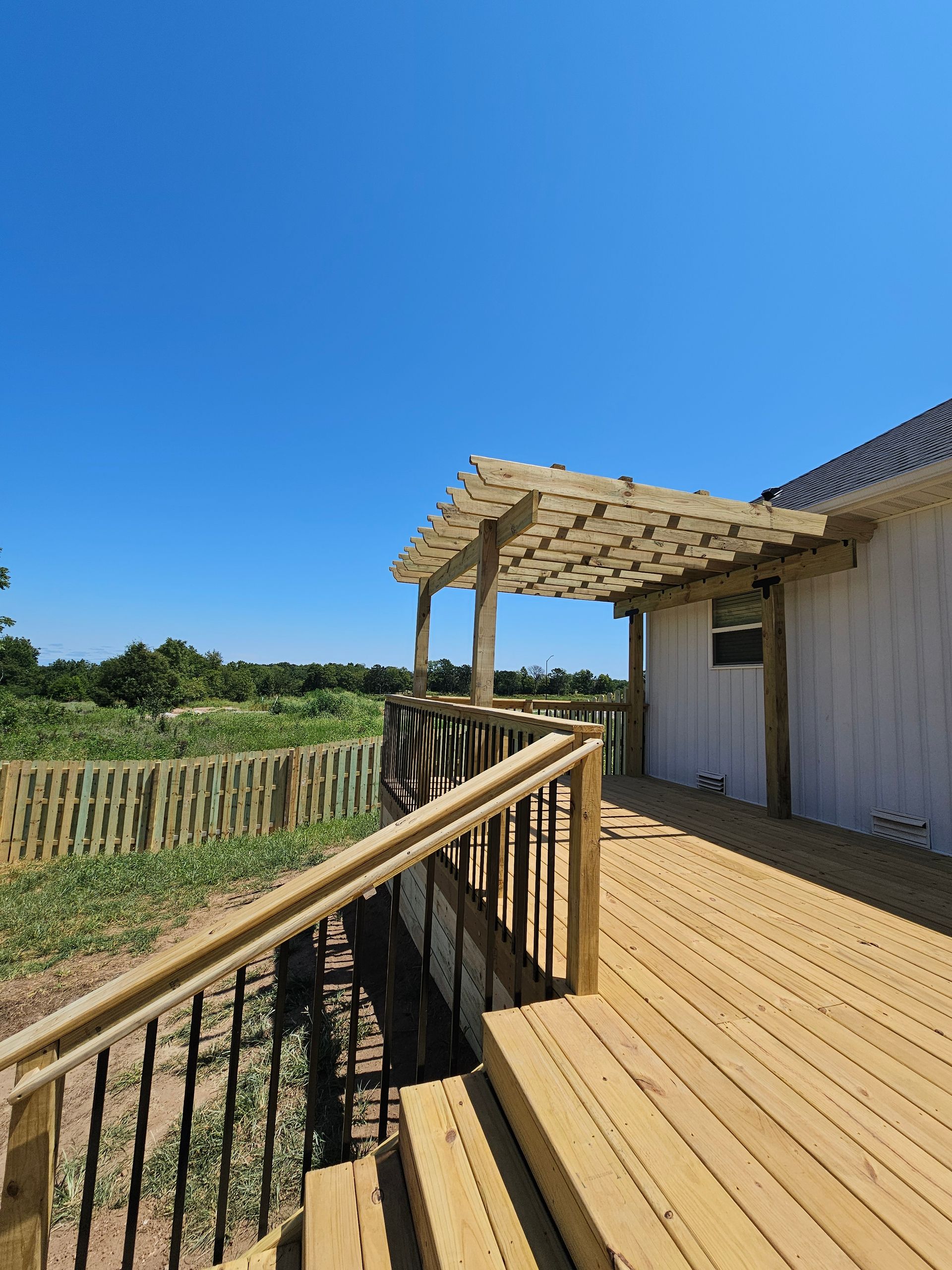Wooden deck with pergola, railing, and fence on a sunny day. Building exterior and blue sky in the background.