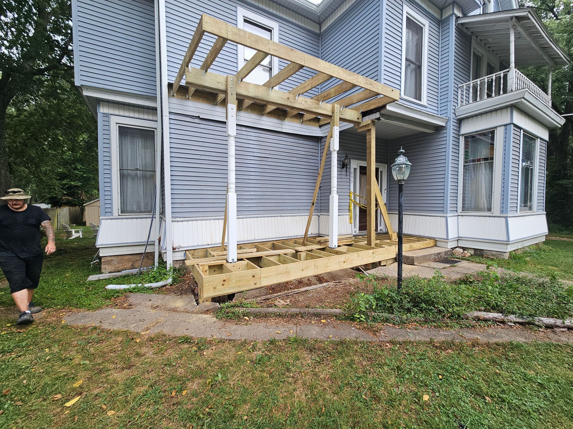 Wooden deck and pergola being built on a light blue house's side. A person walks by.