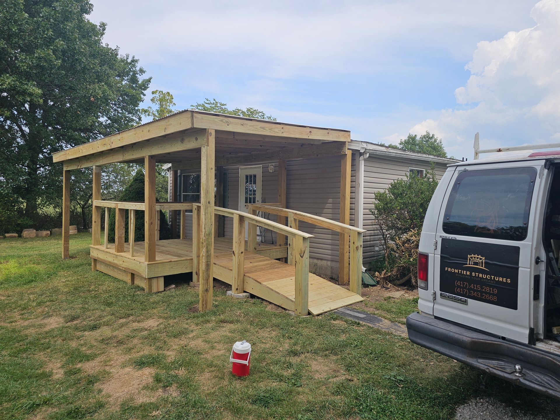 New wooden porch and ramp built onto a small building, outdoors. Includes a covered roof, railings, and a van.