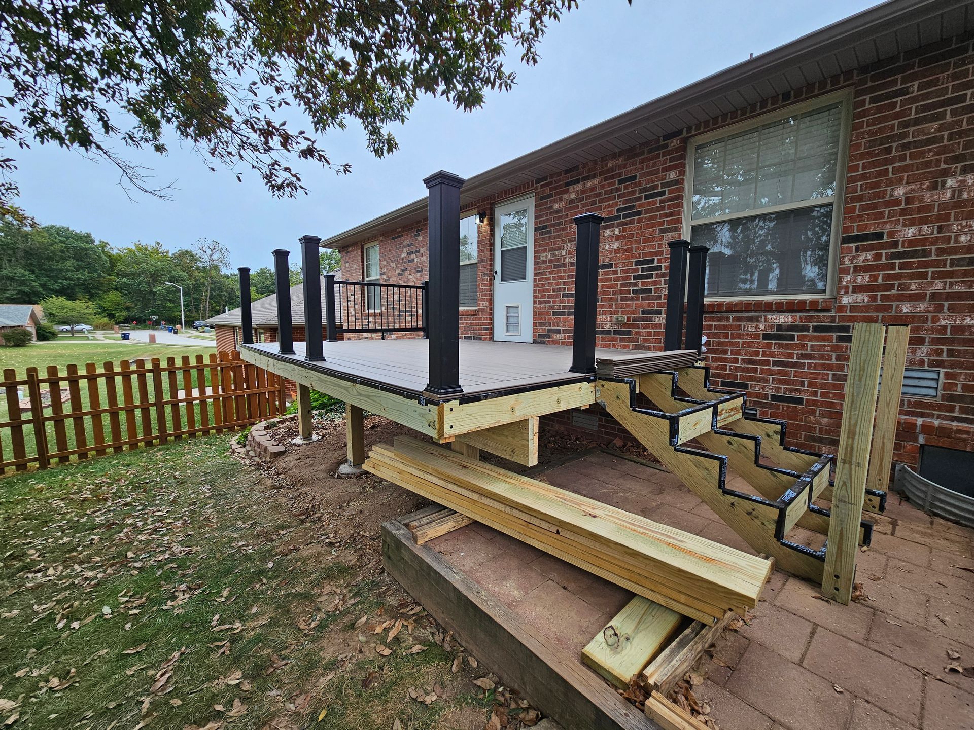 A newly built deck attached to a brick house with stairs, brown posts and railing, and a small fence.
