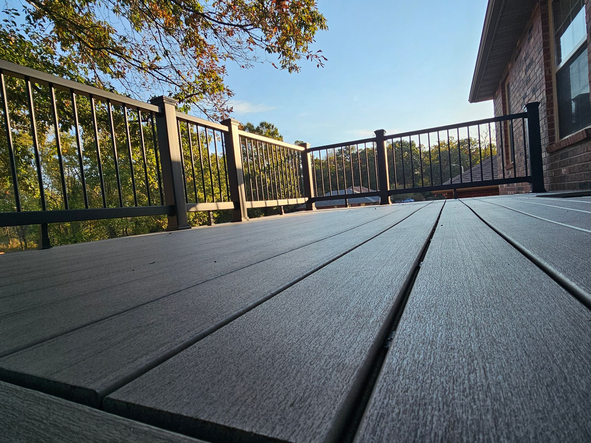 Dark gray composite deck with black metal railing, against a brick house. Trees and blue sky visible.