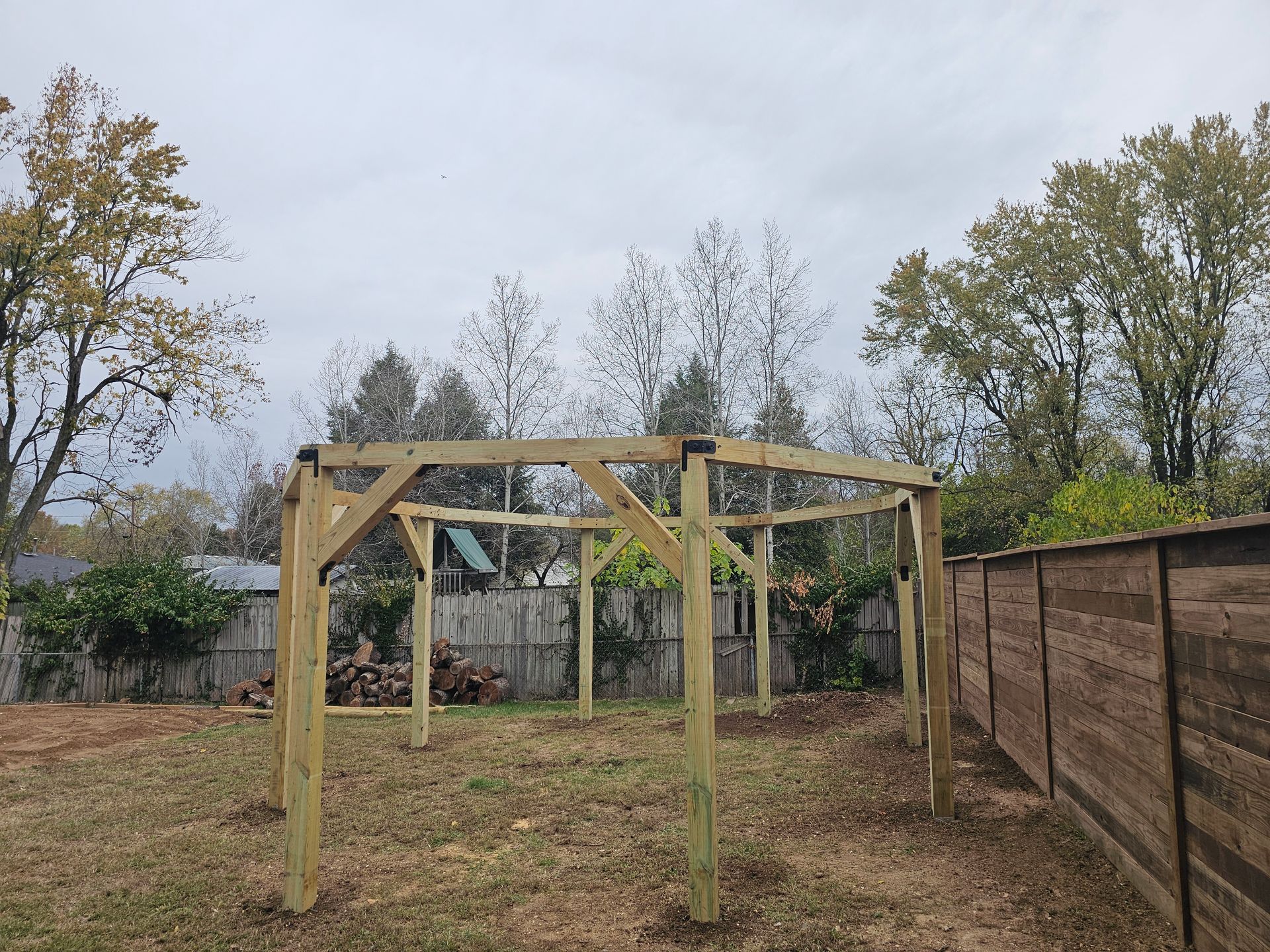 Wooden frame of a gazebo under construction in a grassy backyard, surrounded by trees and a fence.