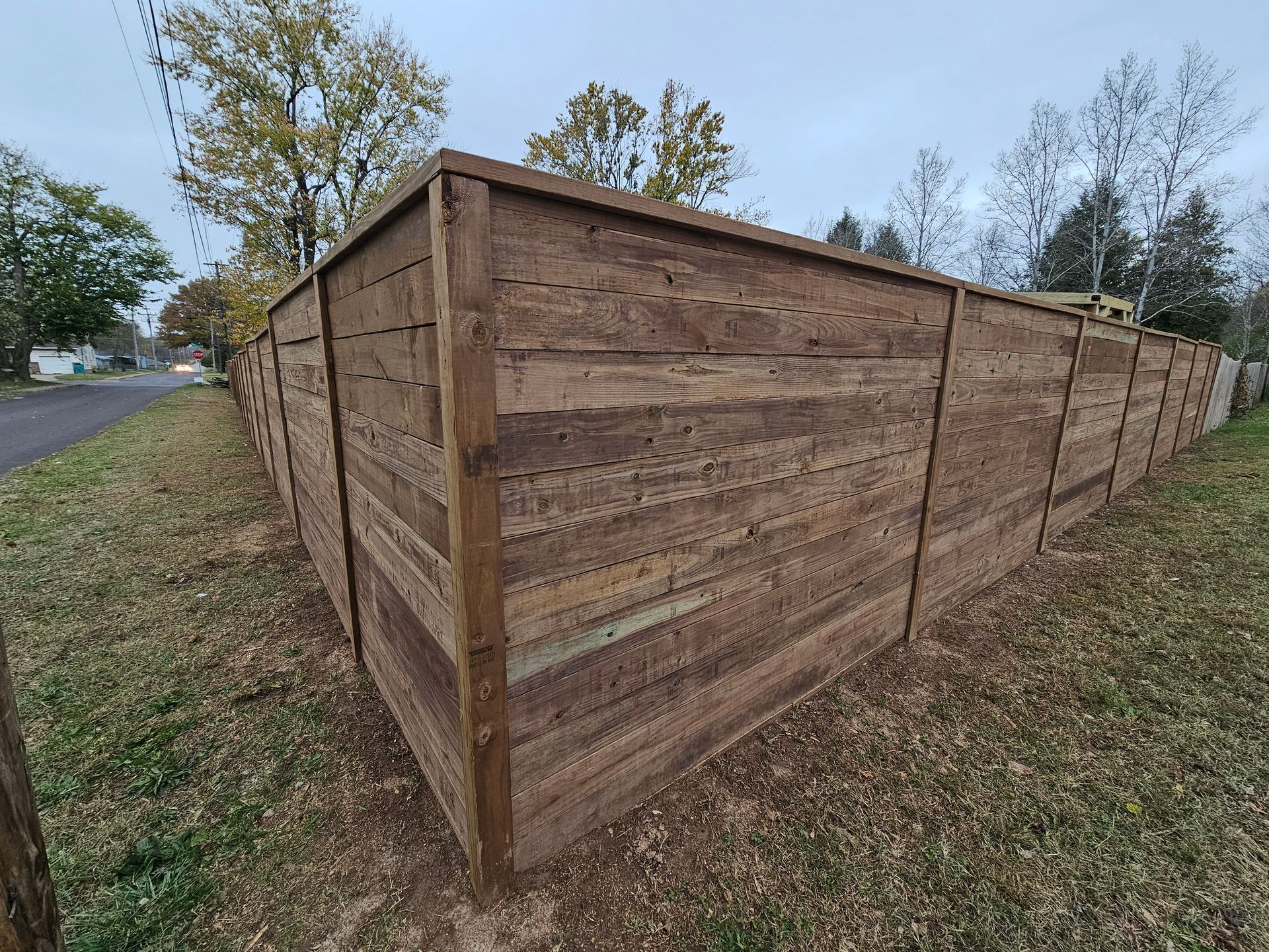 Wooden privacy fence along a grassy area, trees in the background, overcast sky.