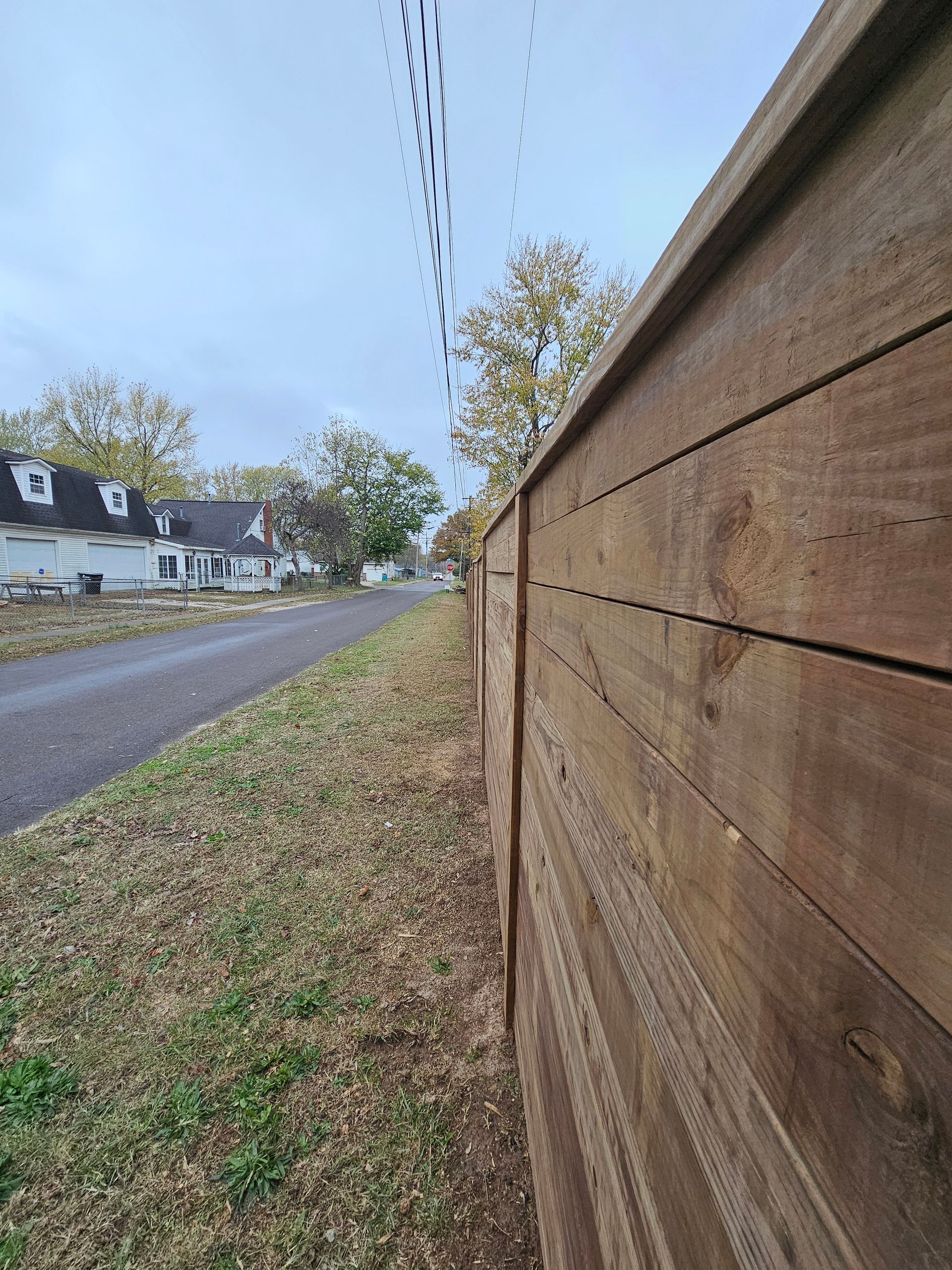 Wooden fence borders a gray path and grassy verge; houses and trees are in the distance under a cloudy sky.