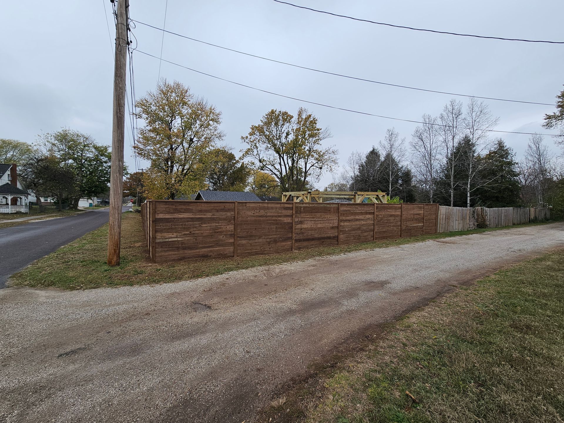 Gravel road leading to a wood fence around a yard with trees under a cloudy sky.