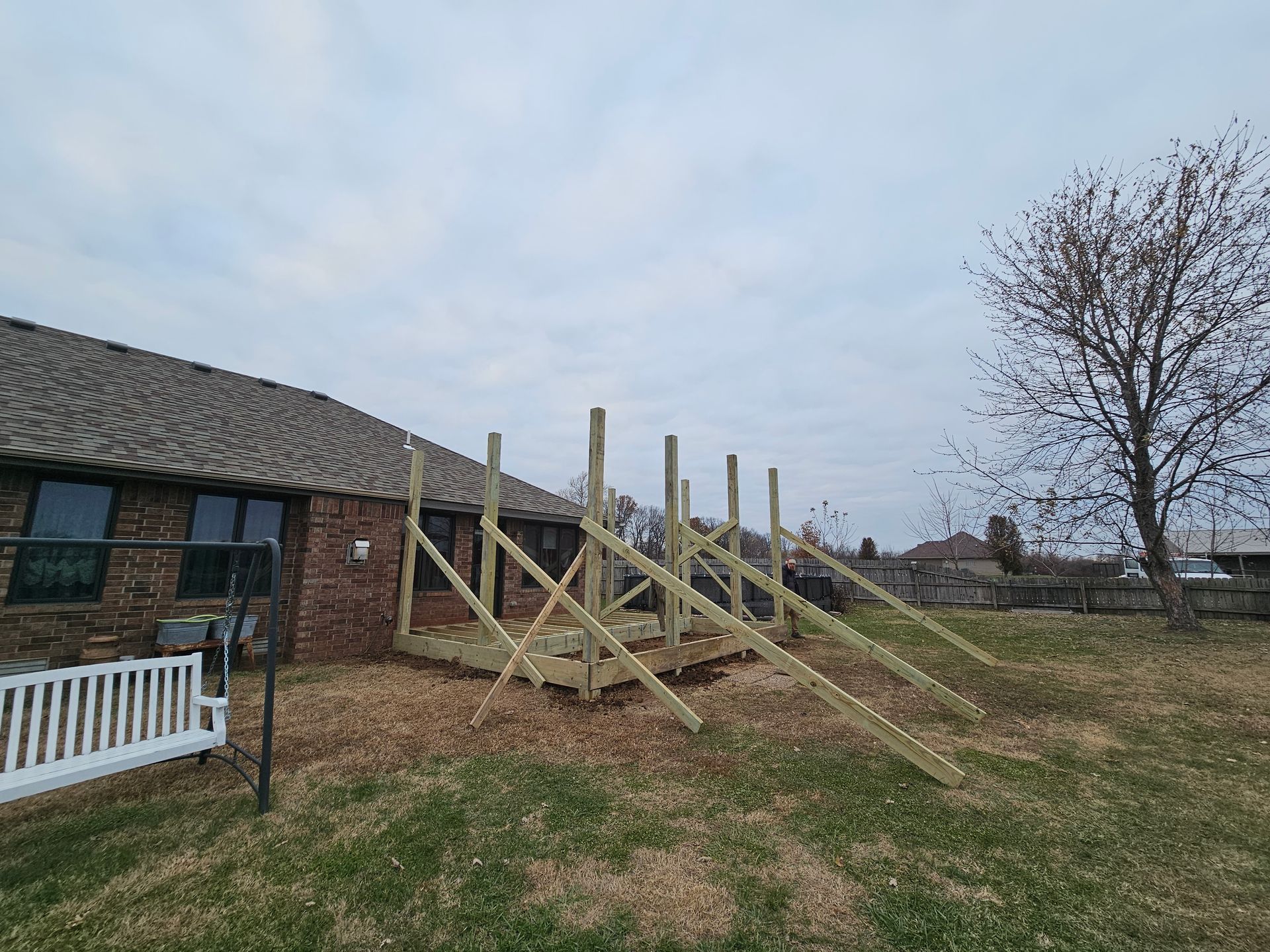 Construction of a deck in a backyard. Wooden frame and posts are partially built. Brown grass and a brick house visible.