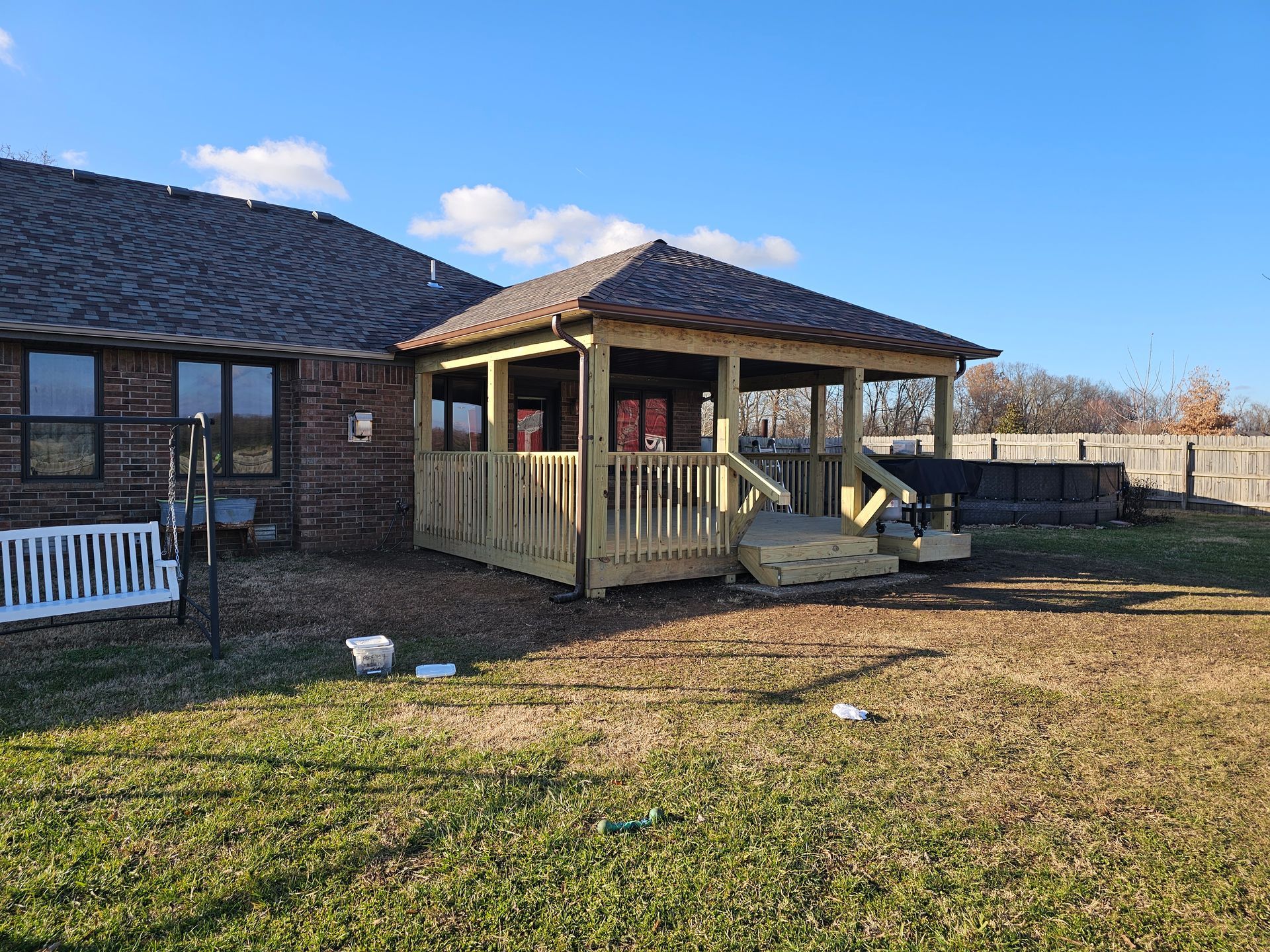 Wooden deck with a covered gazebo, attached to a brick house. Grass and a fence in the background.
