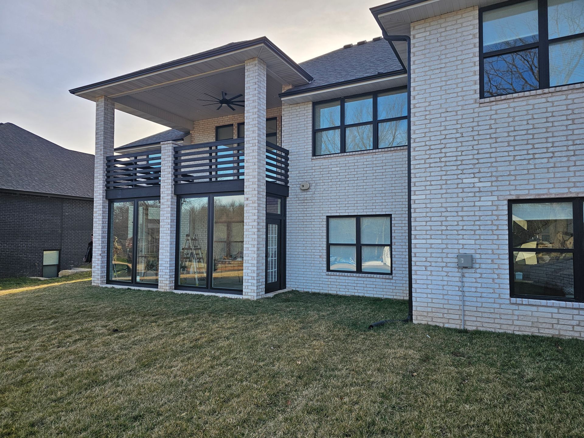 Back of a white brick house with a covered patio, balcony, and dark-framed windows against a clear sky.